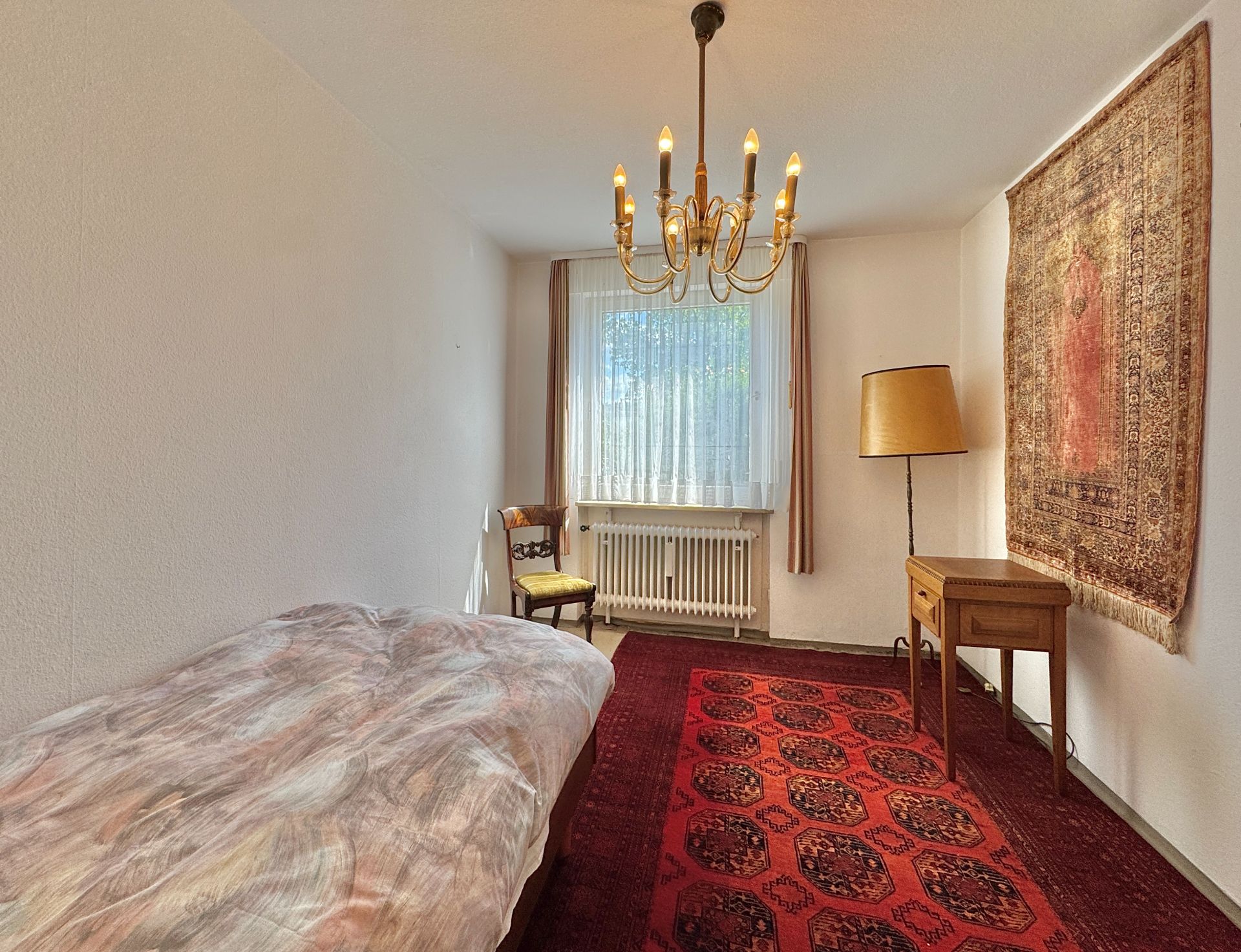 Schlafzimmer mit Bett, Stuhl, Schreibtisch, Stehlampe, Teppich und Wandteppich.
