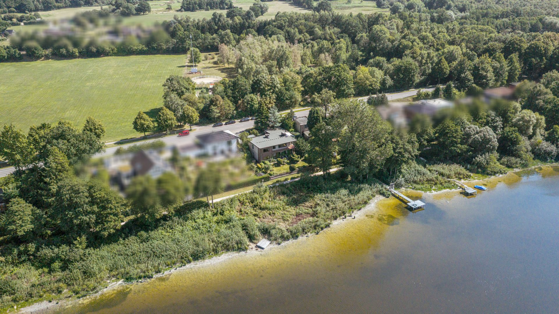 Haus am See mit Steg, umgeben von Bäumen und Wiese, Luftaufnahme.