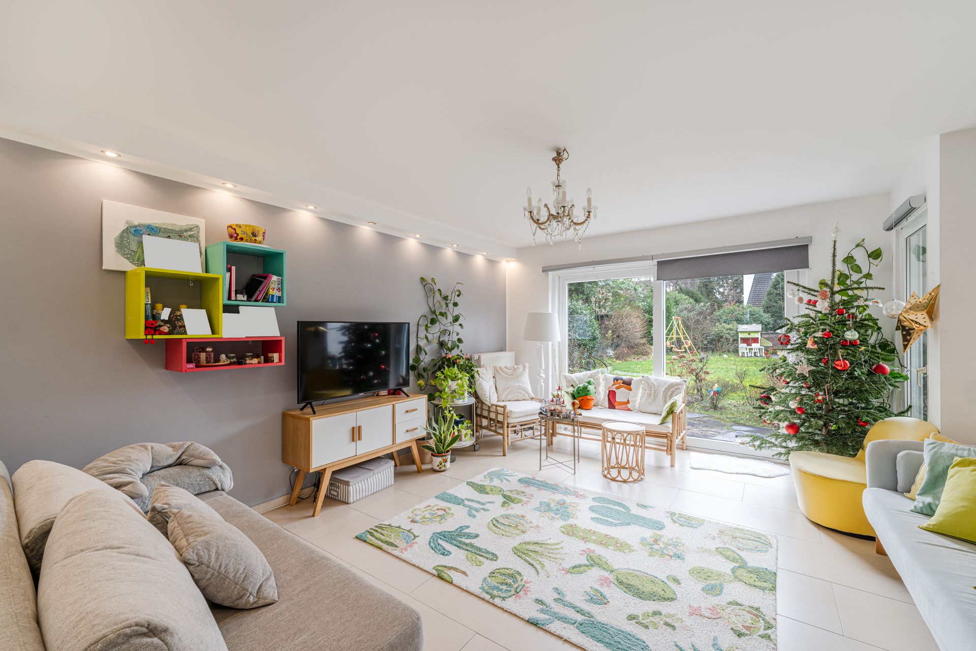 Helles Wohnzimmer mit Sofa, TV, buntem Regal, Weihnachtsbaum und Blick in den Garten.