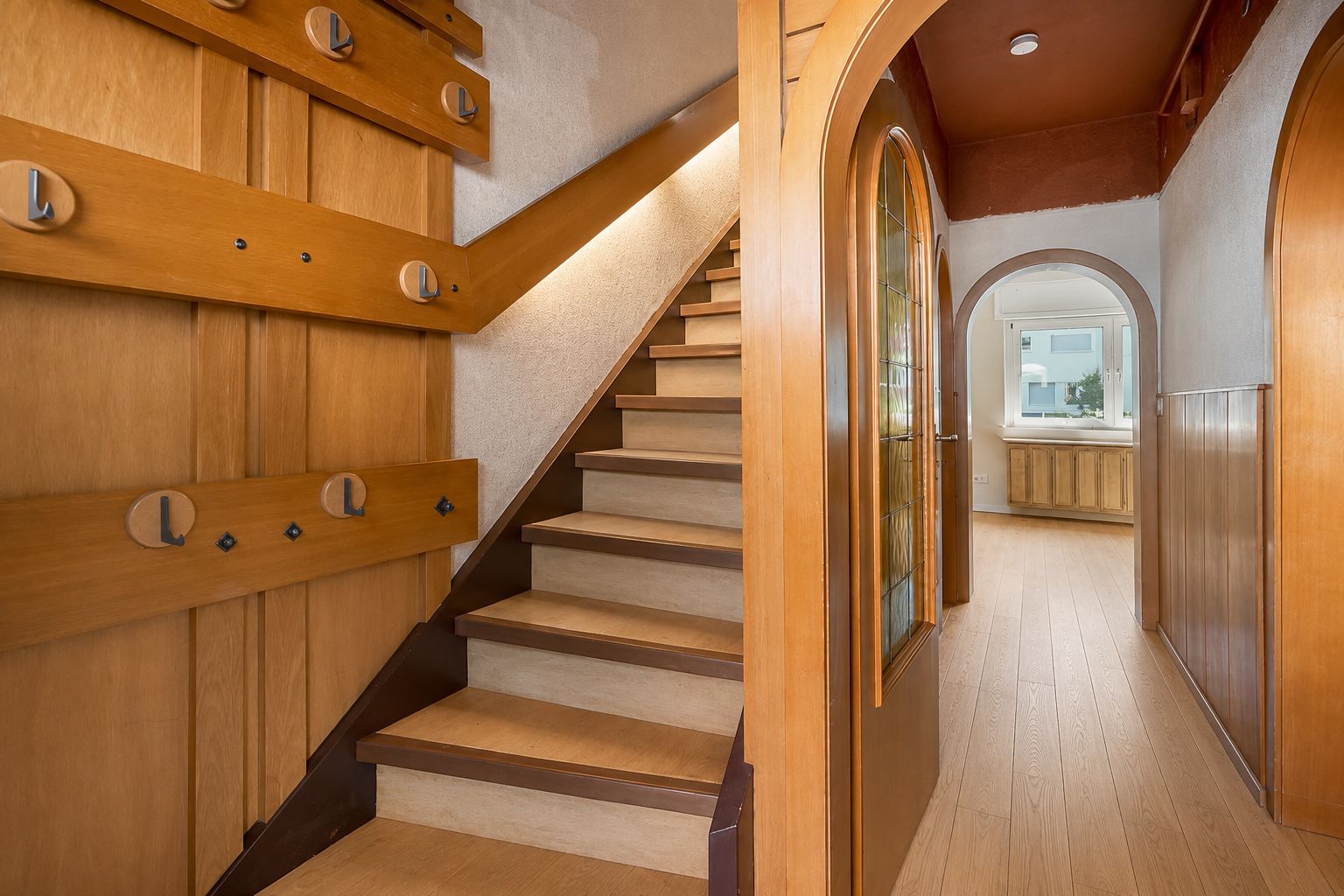 Flur mit Holztreppe, Holzdetails und gewölbtem Durchgang zu einem hellen Raum.