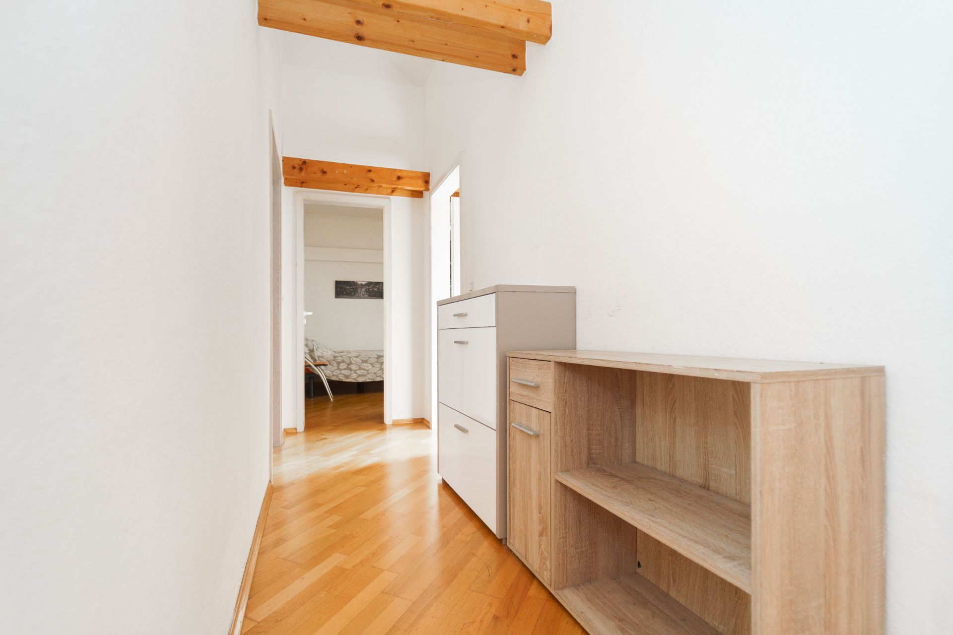 Heller Flur mit Holzboden, Deckenbalken und Holzregal, Blick ins angrenzende Zimmer.