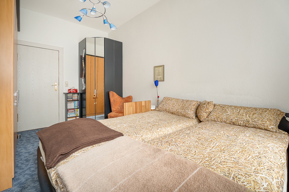 Schlafzimmer mit zwei Einzelbetten, Kleiderschrank, Regal und braunem Sessel.