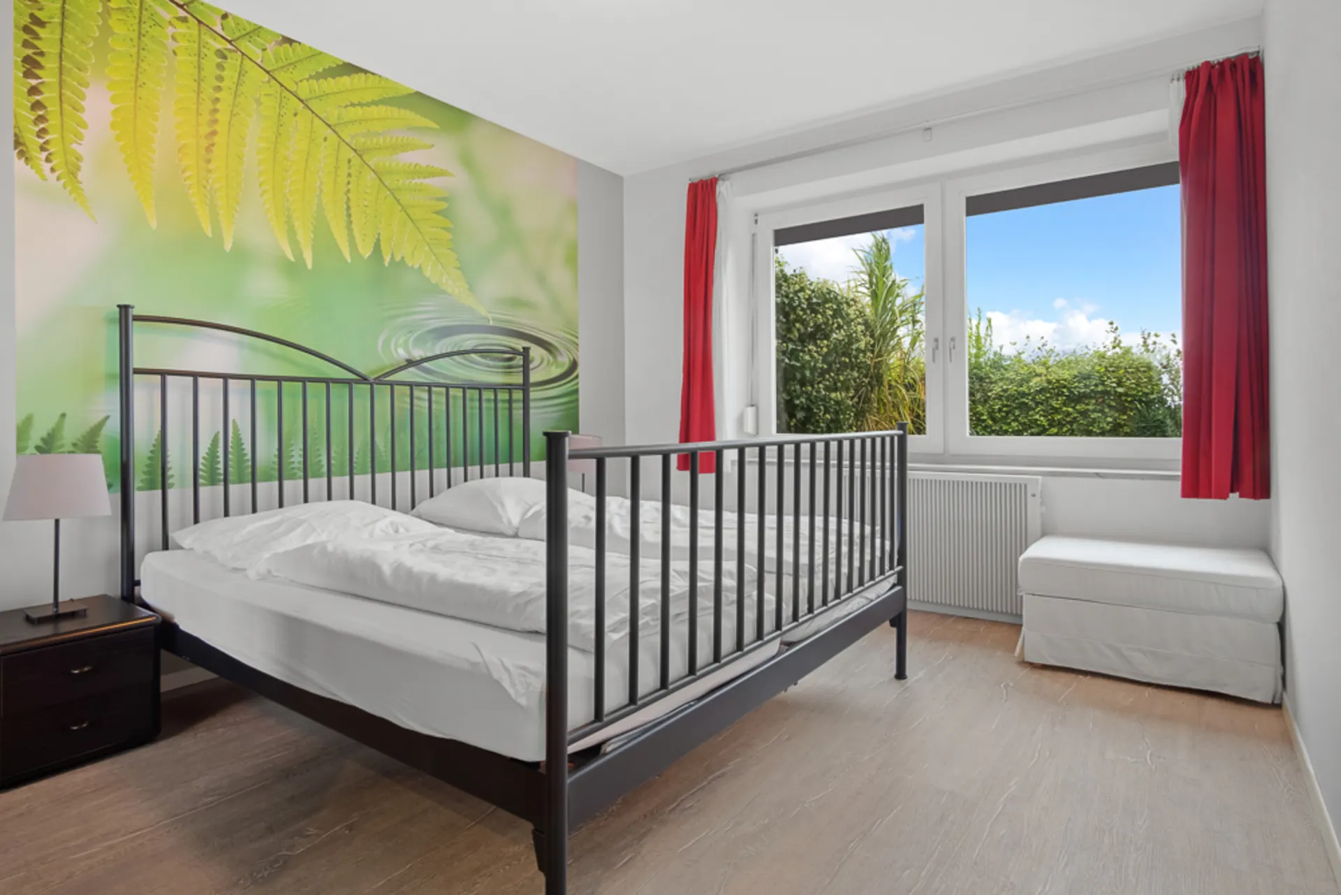 Helles Schlafzimmer mit Metallbett, großem Fenster, roten Vorhängen und Wandbild mit Farn.