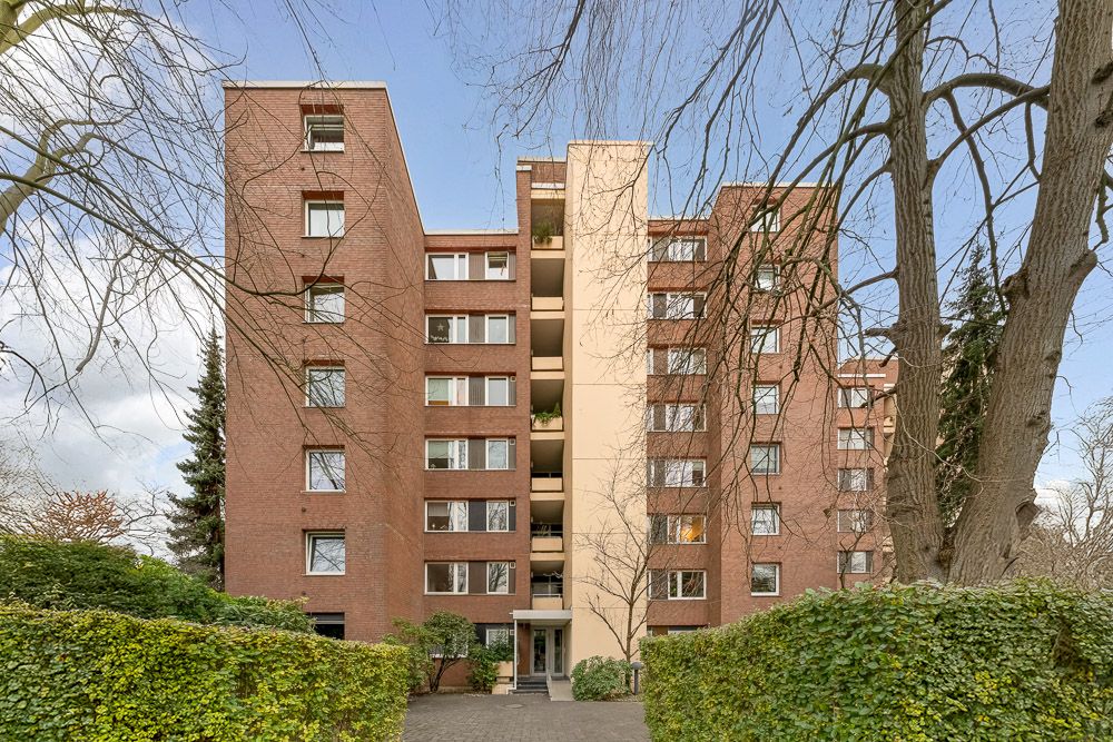 Mehrstöckiges Wohnhaus aus rotem Backstein mit Eingangsbereich, umgeben von Bäumen und Hecken.