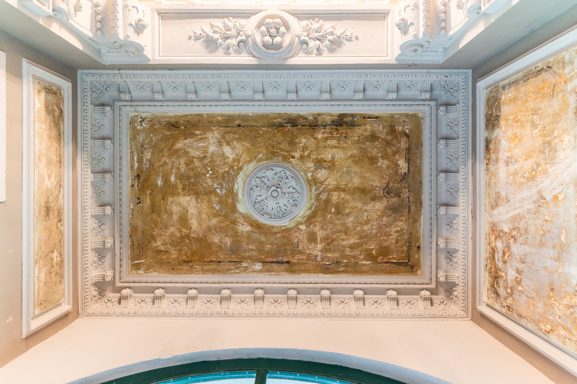 Verzierte, gealterte Decke mit Stuckrahmen und einem runden Ornament in der Mitte.