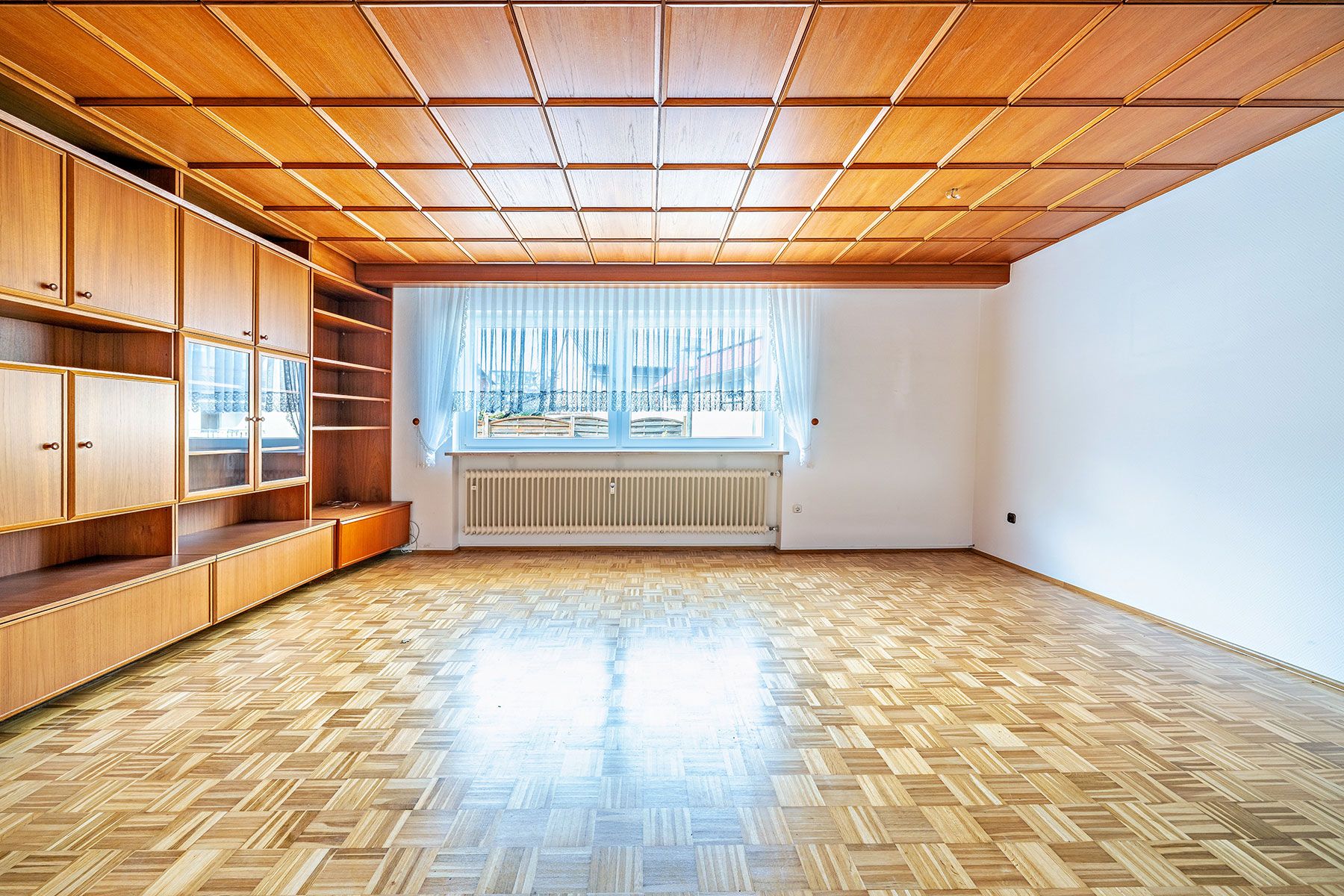 Helles Wohnzimmer mit Holzdecke, Parkettboden, Einbauschrank und großem Fenster mit Vorhang.