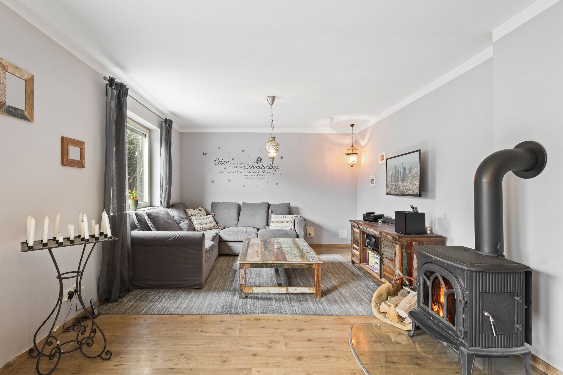 Helles Wohnzimmer mit grauem Sofa, Holztisch, Kaminofen und Wanddeko.