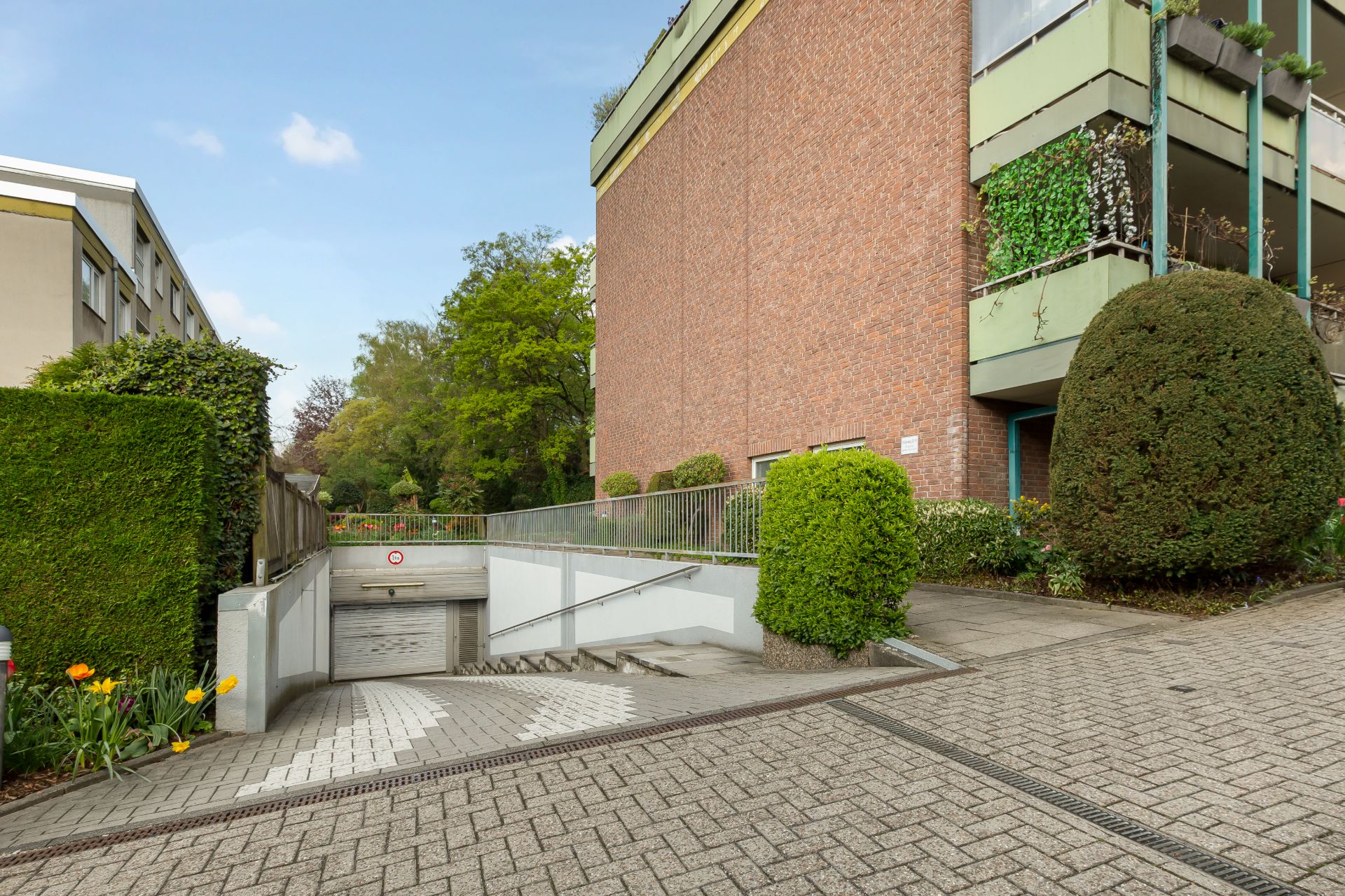 Einfahrt zu einer Tiefgarage mit Treppe und gepflegten Büschen neben einem Wohnhaus.
