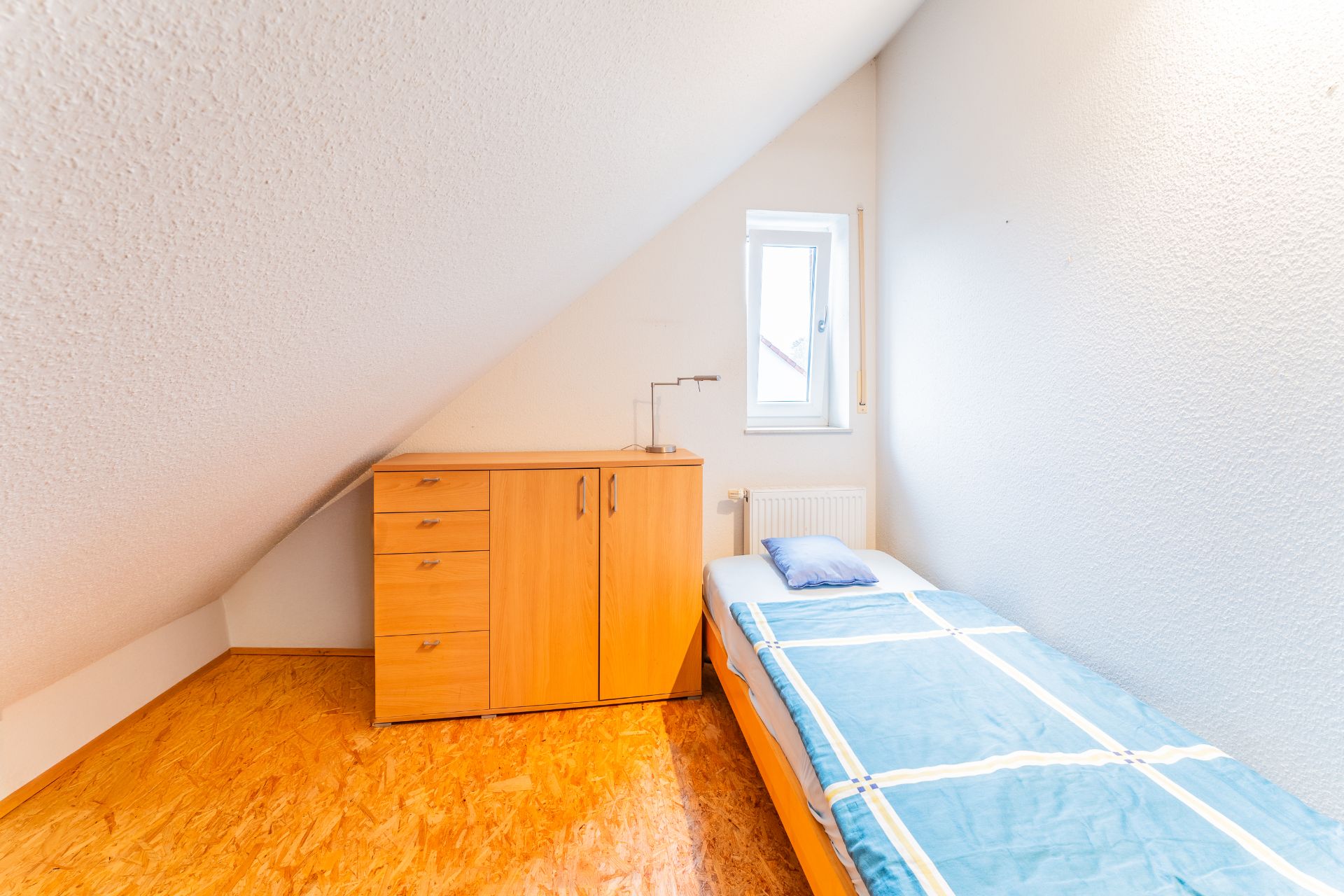 Kleines Zimmer mit Einzelbett, Holzboden, Kommode und Fenster unter Dachschräge.