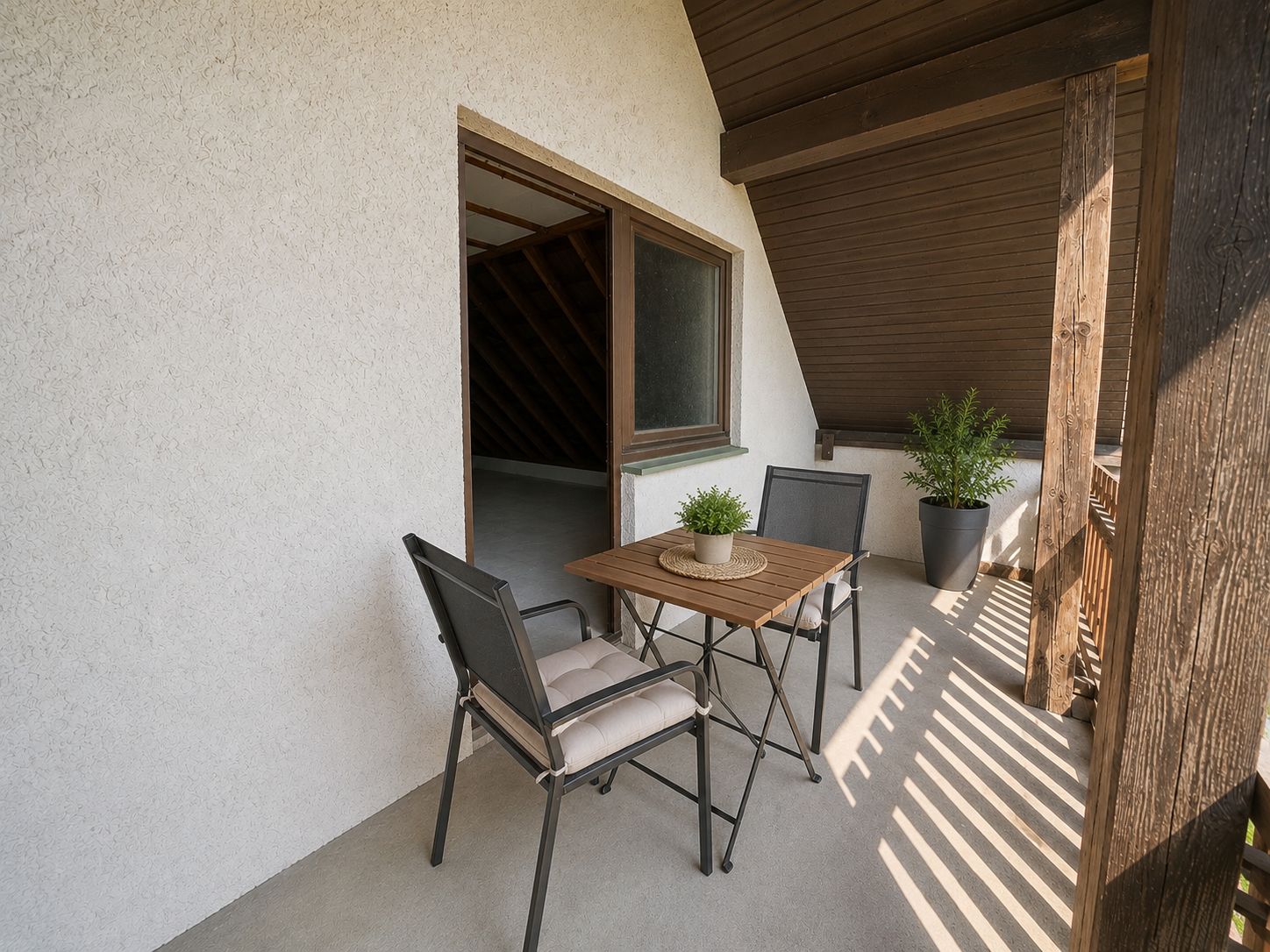 Überdachter Balkon mit kleinem Tisch, zwei Stühlen und einer Pflanze in der Ecke.