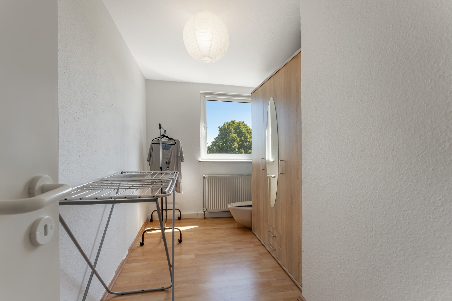Kleines Zimmer mit Kleiderschrank, Wäscheständer und Fenster mit Blick ins Grüne.