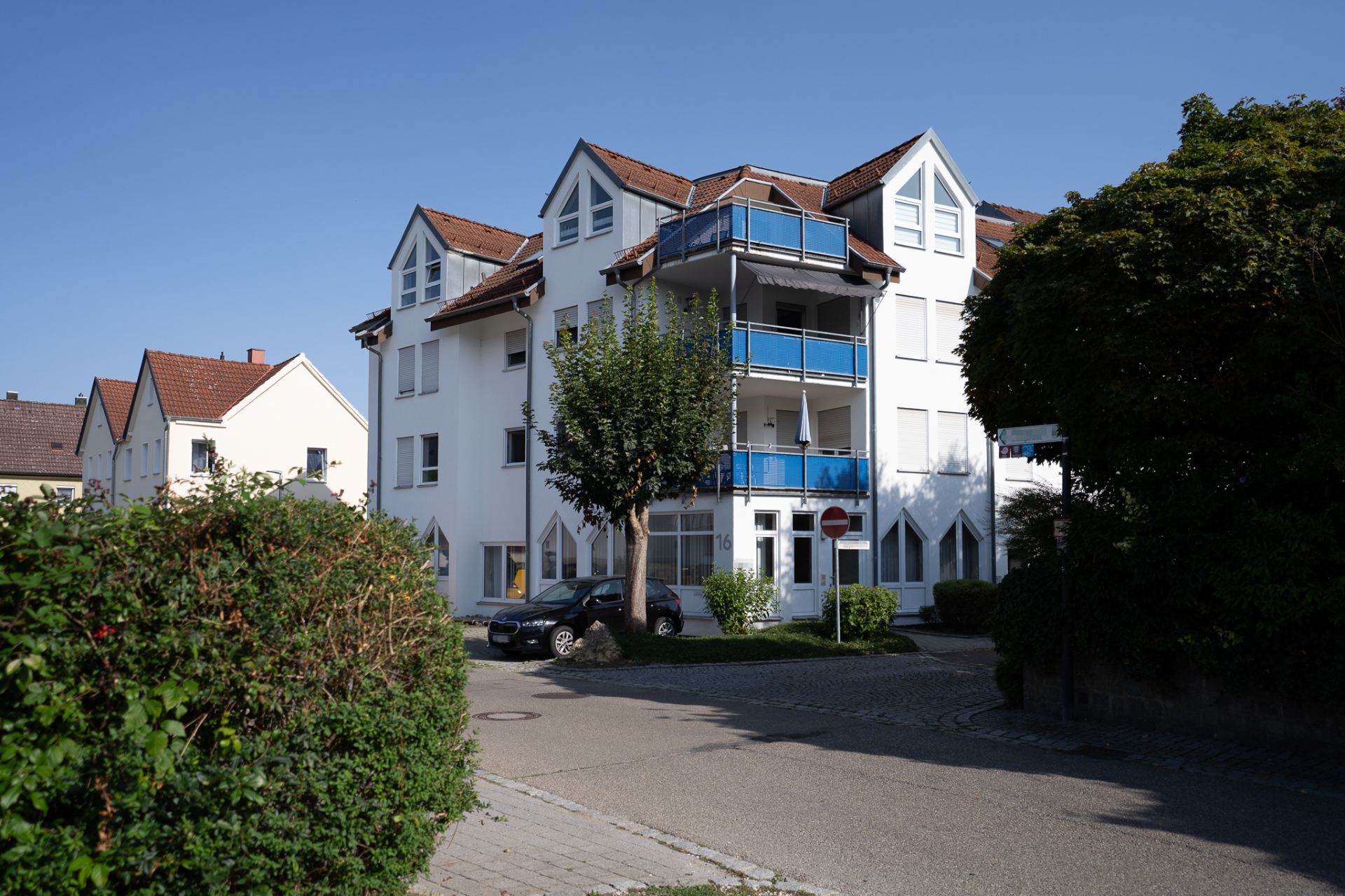 Mehrfamilienhaus mit blauen Balkonen, Bäumen und geparkten Autos bei Sonnenschein.
