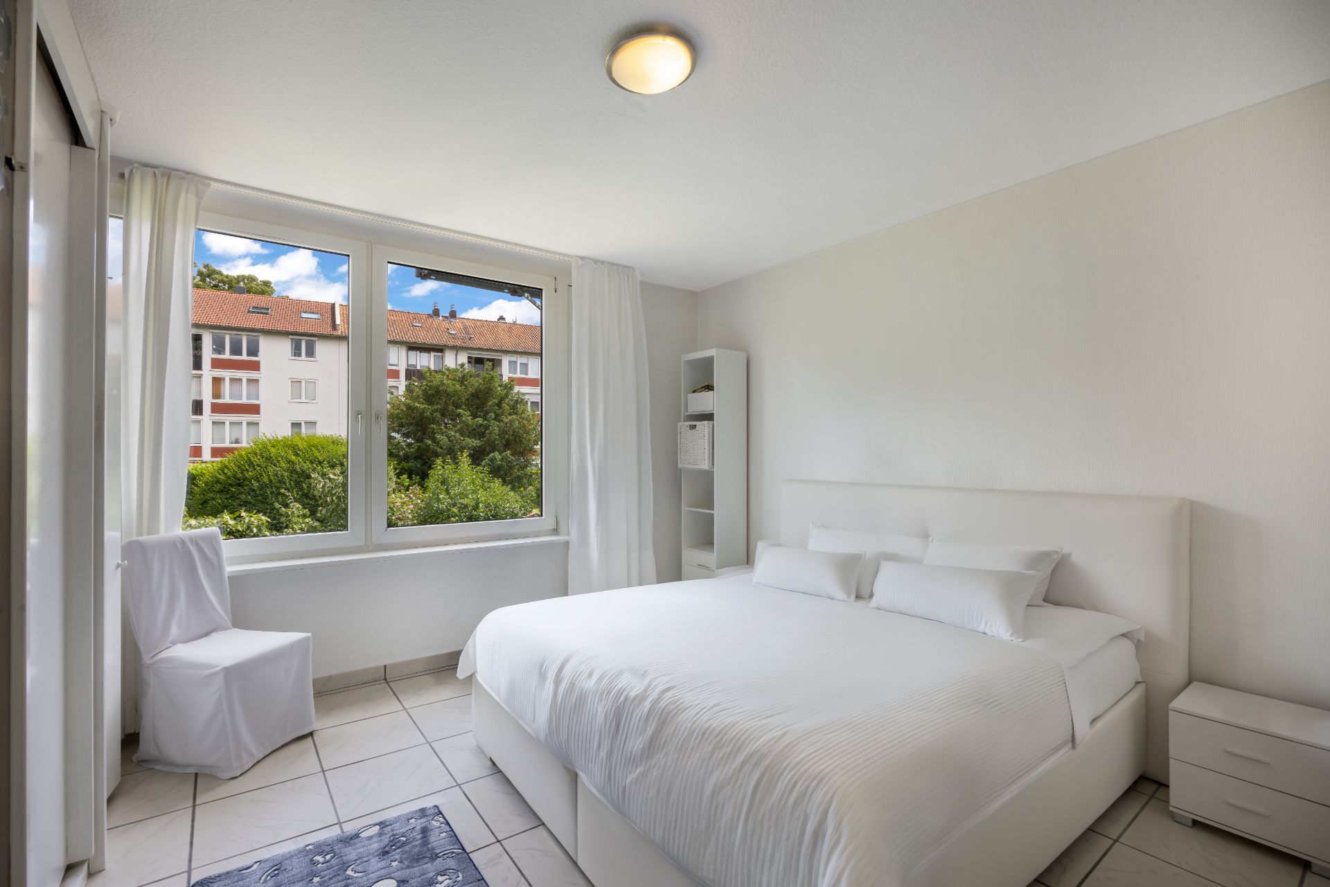 Helles Schlafzimmer mit großem Bett, Stuhl, Fenster und Blick auf grüne Bäume und Nachbargebäude.