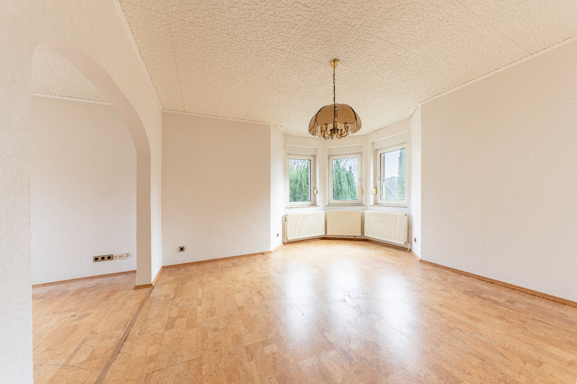 Helles, leeres Zimmer mit Holzfußboden, drei Fenstern, Heizkörpern und einem Deckenleuchter.