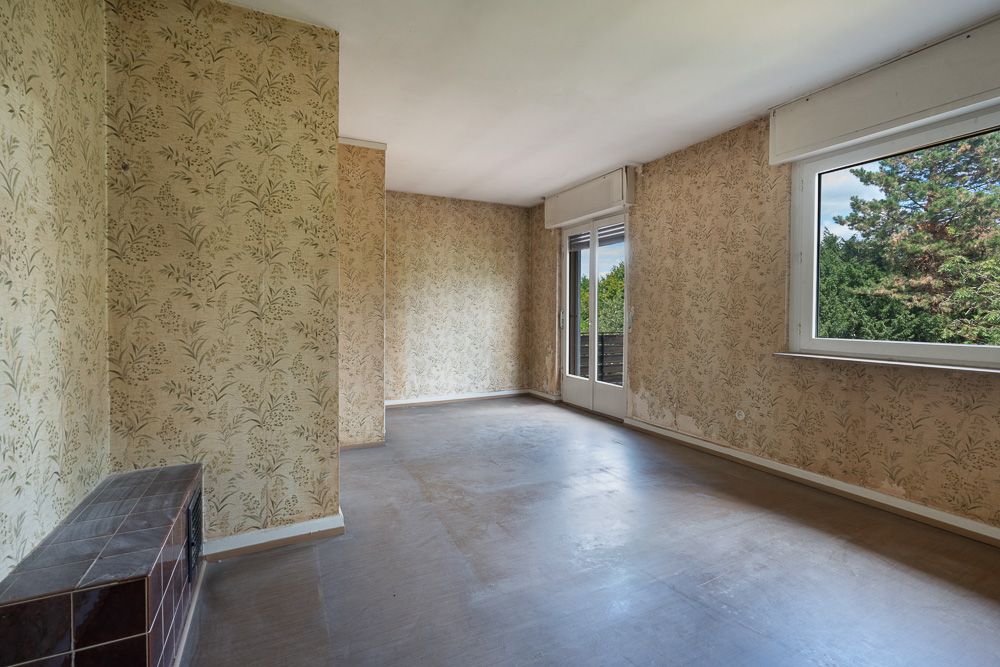 Leeres Zimmer mit gemusterten Tapeten, großem Fenster und Tür zum Balkon, Blick ins Grüne.