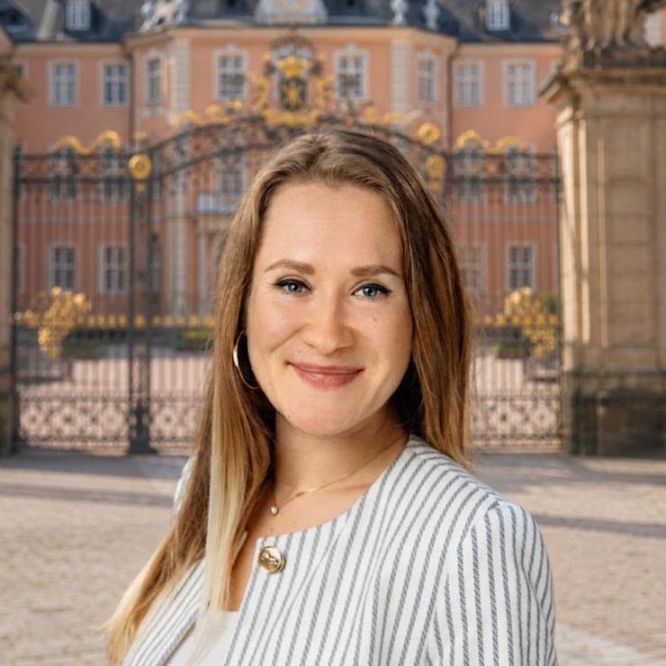 Frau mit gestreifter Jacke steht auf einem gepflasterten Platz vor einem großen Gebäude mit Tor.