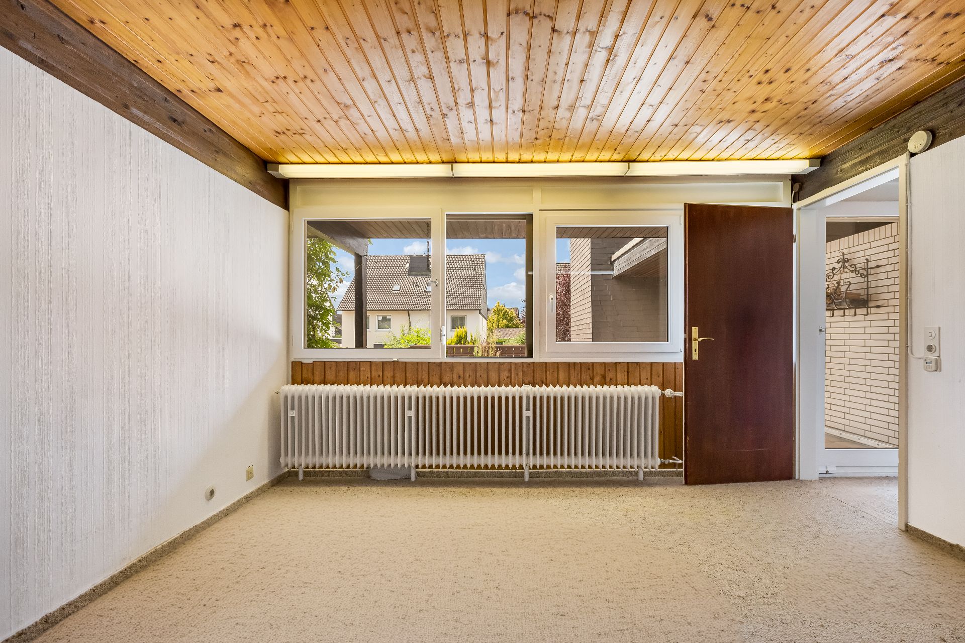 Leerer Raum mit Holzdecke, Teppichboden, Heizkörper und Fensterfront mit geöffneter Tür.