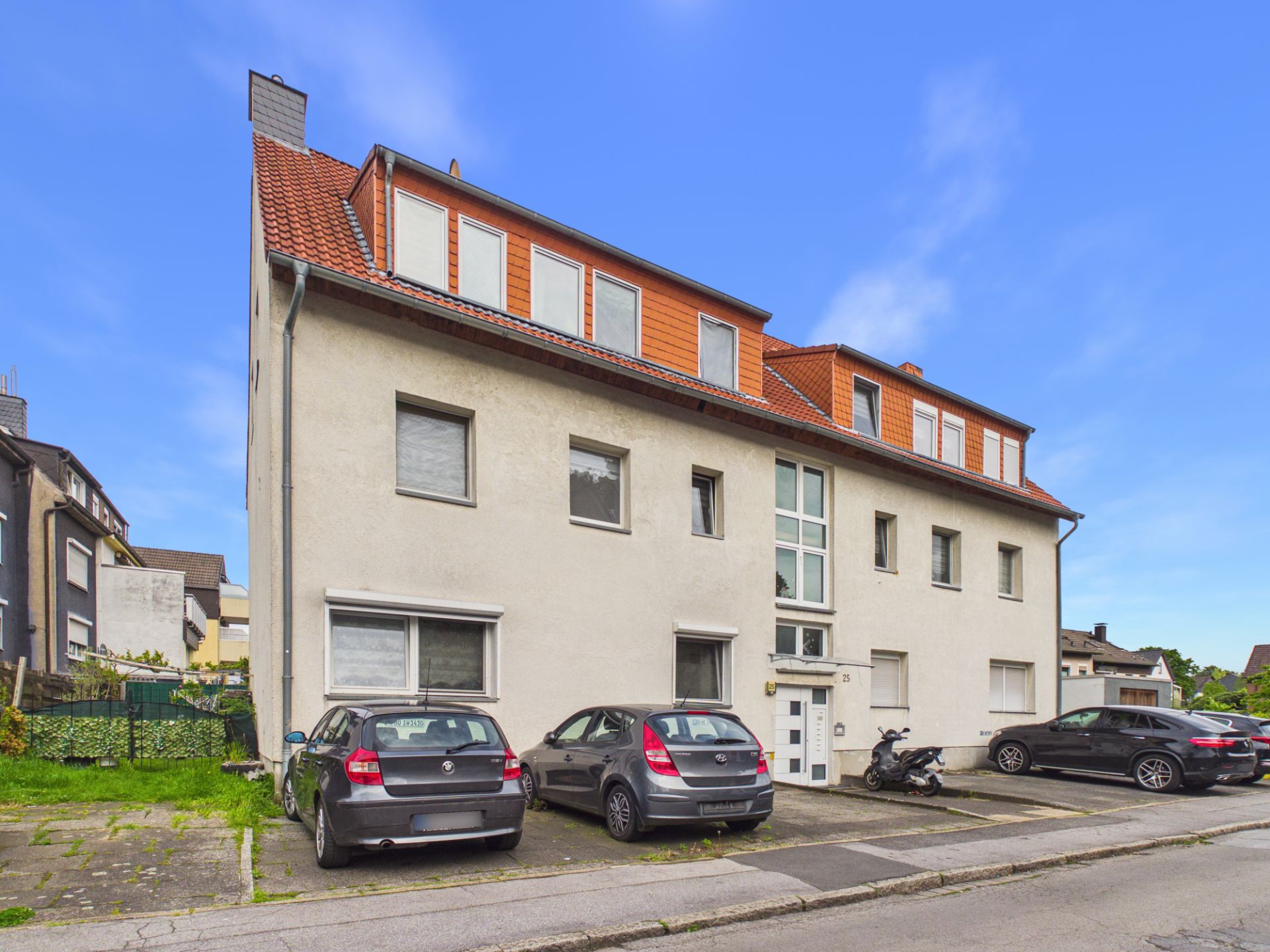 Mehrfamilienhaus mit roten Dachziegeln und mehreren geparkten Autos davor.