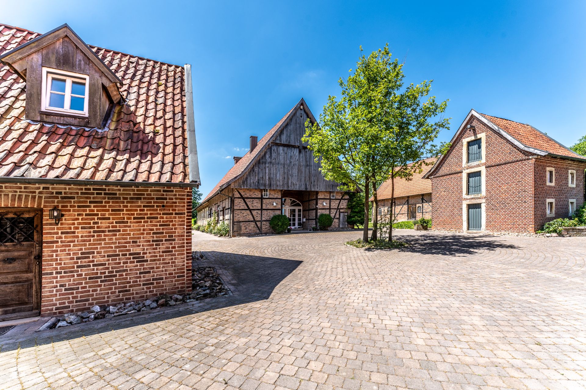 Gepflasterter Hof mit mehreren Backstein- und Fachwerkhäusern sowie einem Baum in der Mitte.
