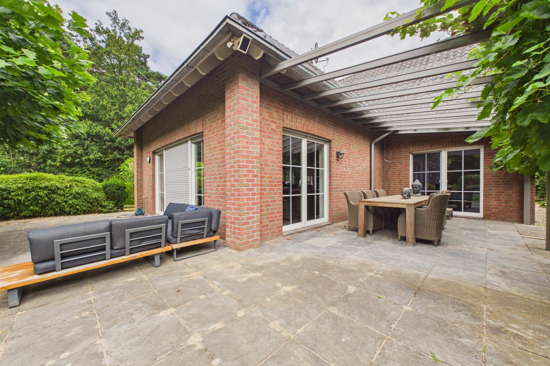 Große Terrasse mit Sitzgruppe und Esstisch, Haus mit roten Ziegelwänden und großen Fenstern.