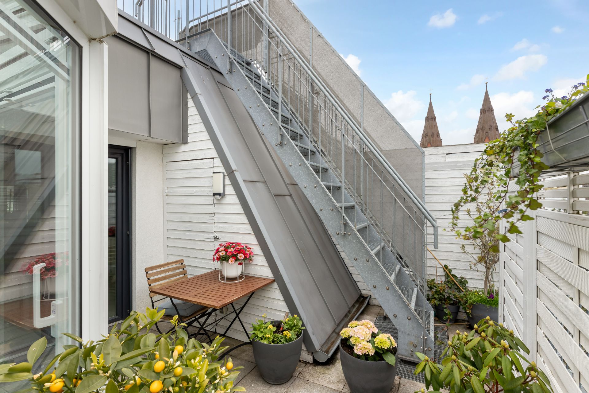 Kleine Terrasse mit Holztisch, Stühlen, Blumentöpfen und Metalltreppe.