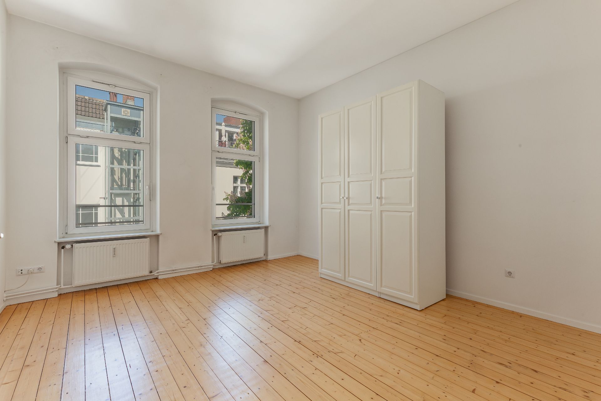 Heller Raum mit Holzfußboden, zwei Fenstern und einem weißen Kleiderschrank.