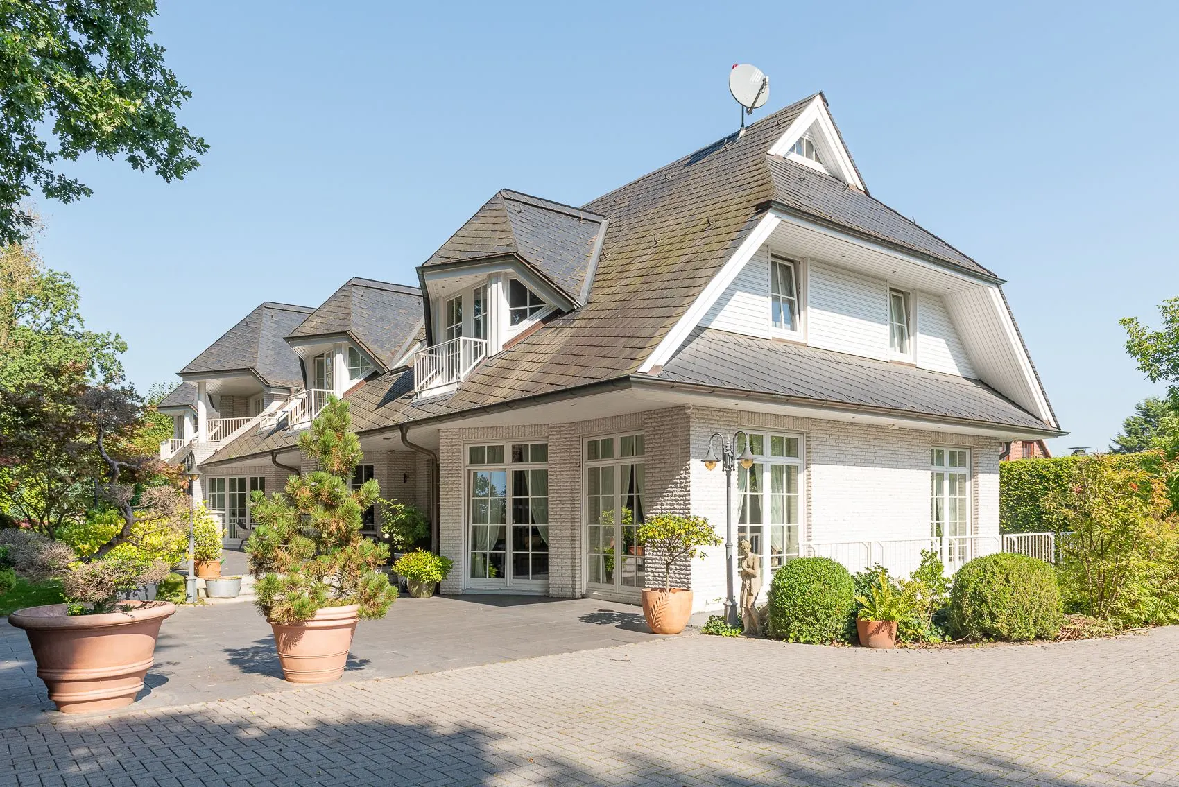 Großes weißes Wohnhaus mit dunklem Dach, vielen Fenstern und Pflanzen im Vorgarten.