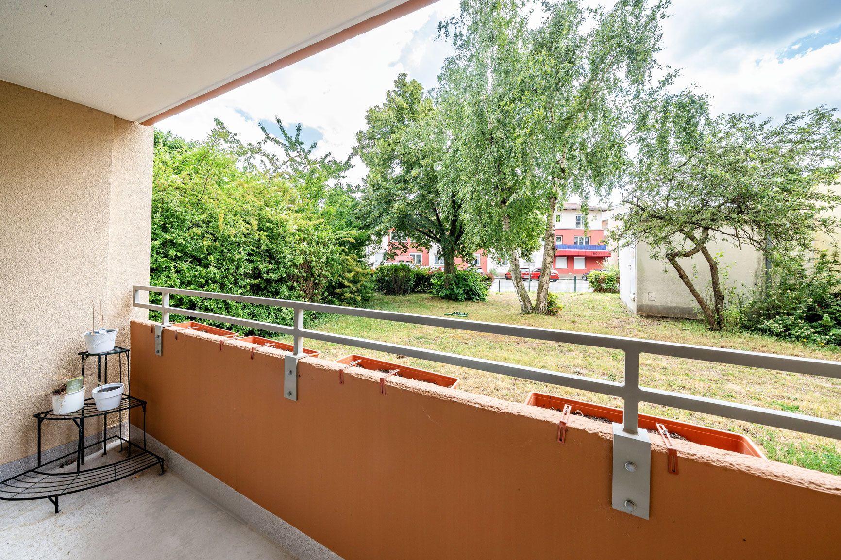 Balkon mit Geländer, Blick auf grünen Garten und Bäume, links Pflanzenregal mit Töpfen.