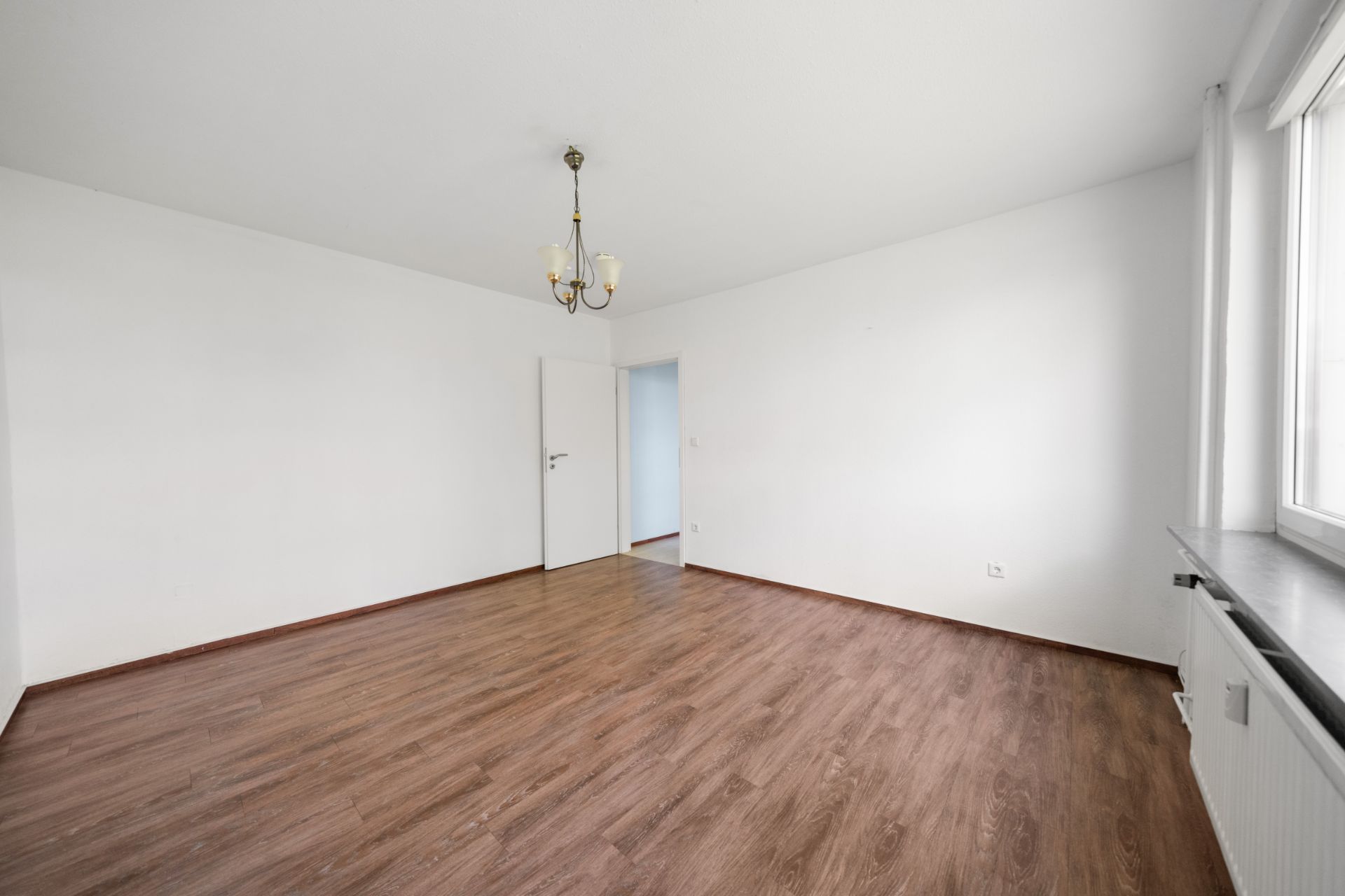 Helles, leeres Zimmer mit Holzfußboden, weißer Wand und Deckenleuchte.