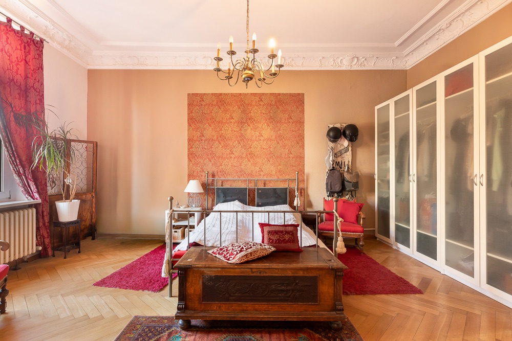 Großes Schlafzimmer mit Bett, Kronleuchter, Holztruhe, Kleiderschrank und roten Akzenten.