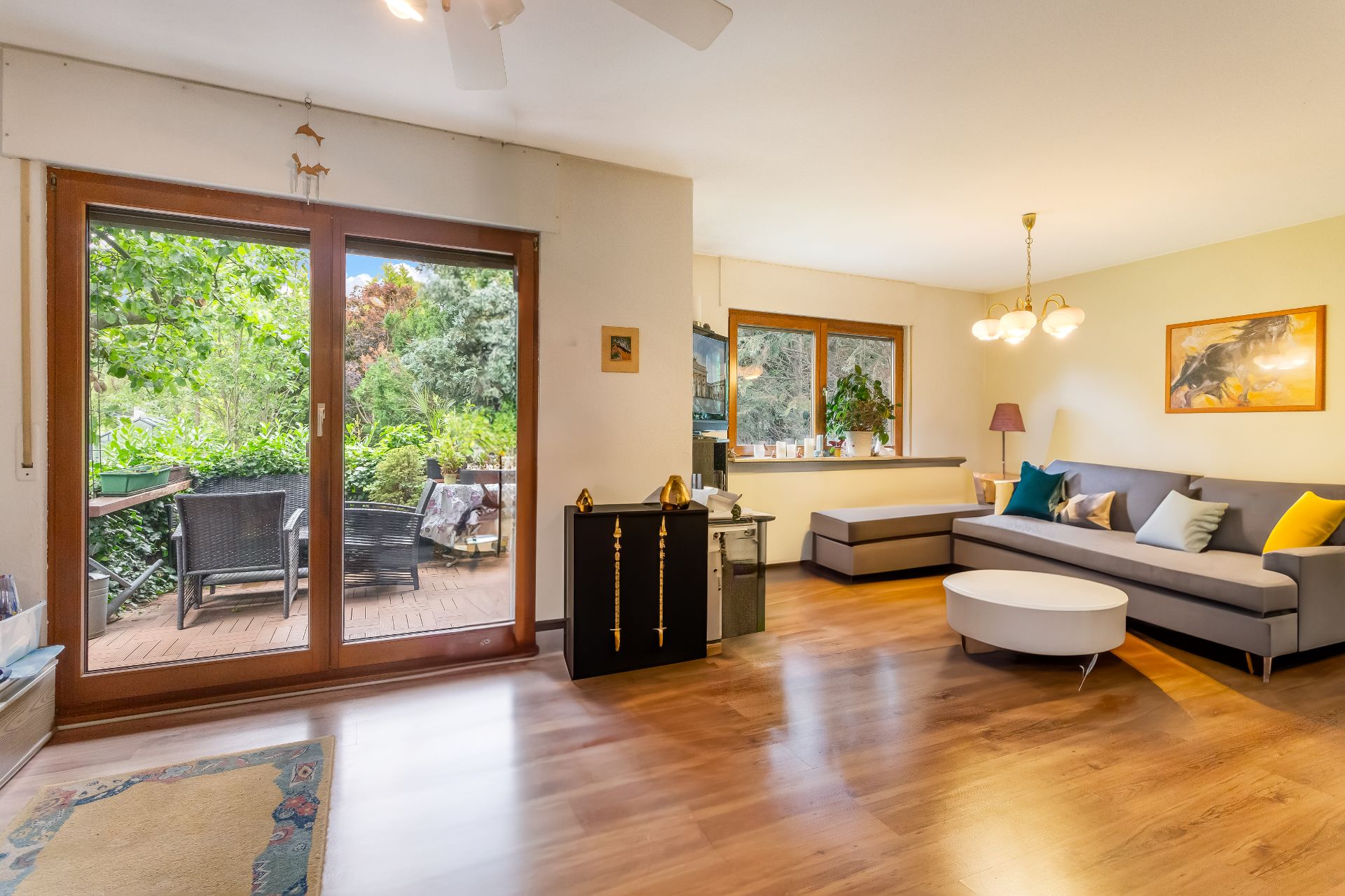 Helles Wohnzimmer mit Ecksofa, rundem Tisch und Zugang zur Terrasse mit Gartenmöbeln.