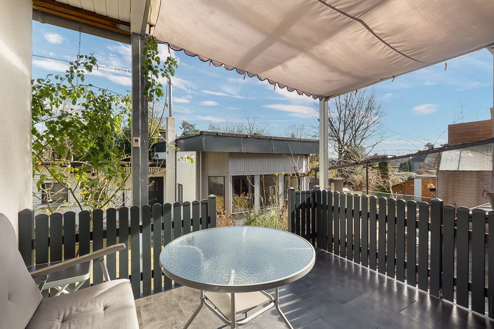 Überdachte Terrasse mit rundem Glastisch, Sofa und schwarzem Zaun, Blick auf den Garten.