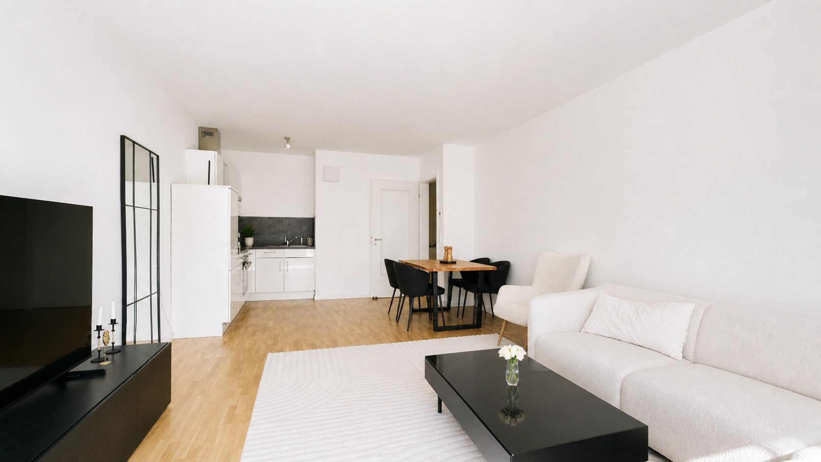 Helles Wohnzimmer mit weißem Sofa, schwarzem Tisch, Esstisch, Fernseher und offener Küche.