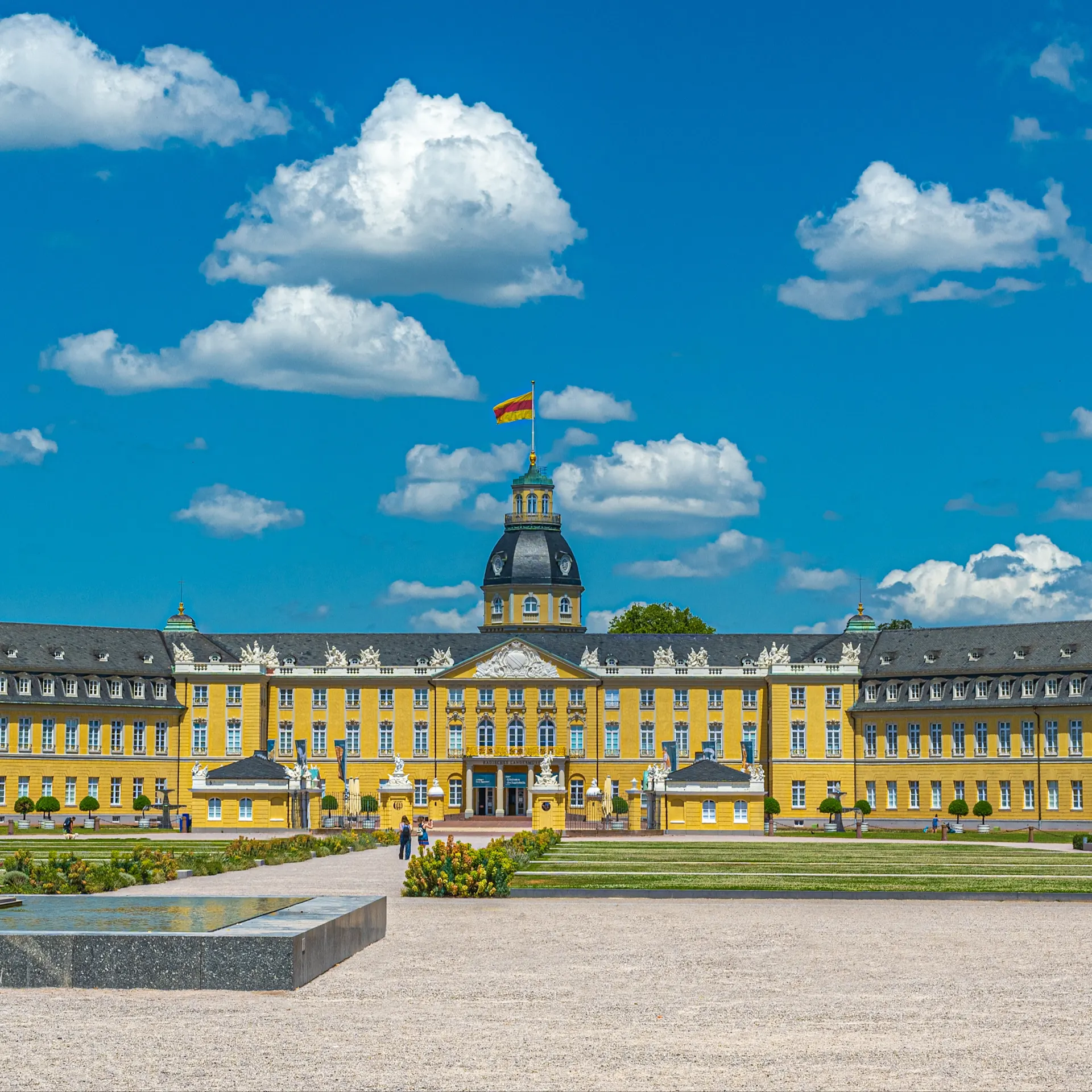 Großes gelbes Schloss mit Turm, Flagge und gepflegtem Garten im Vordergrund.