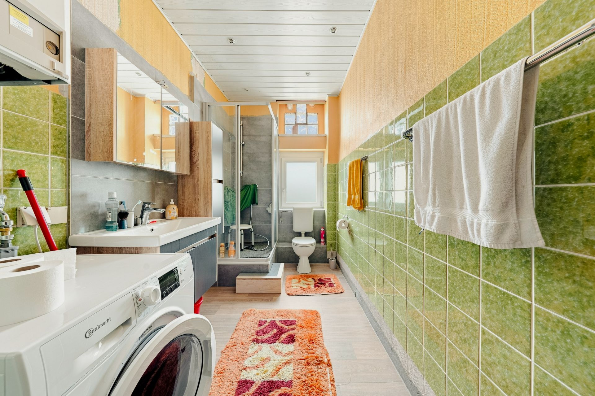 Helles Badezimmer mit grünen Fliesen, Waschmaschine, Dusche, WC und Waschbecken.