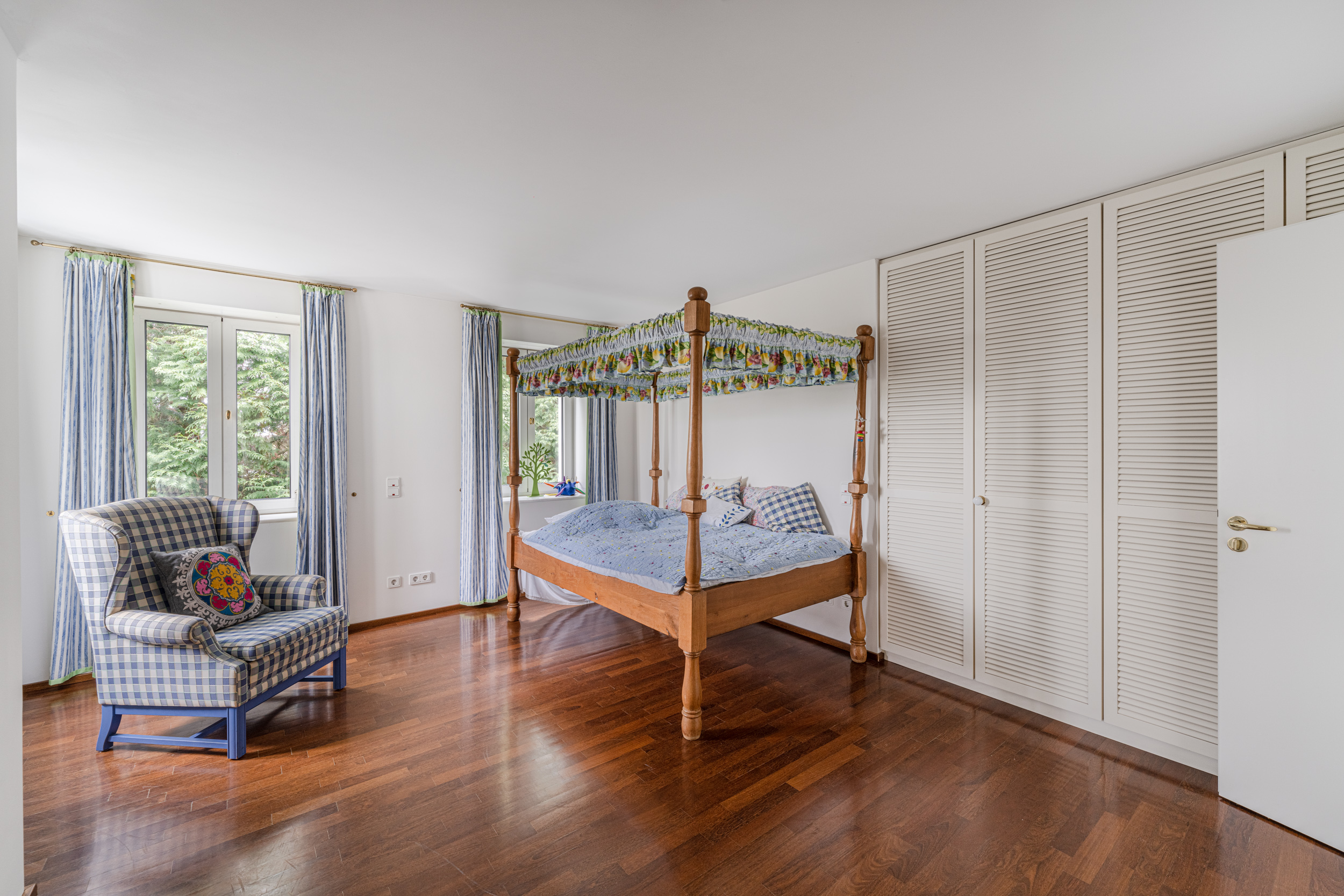 Helles Schlafzimmer mit Himmelbett, Sessel, Holzfußboden und Fenstern mit blauen Vorhängen.