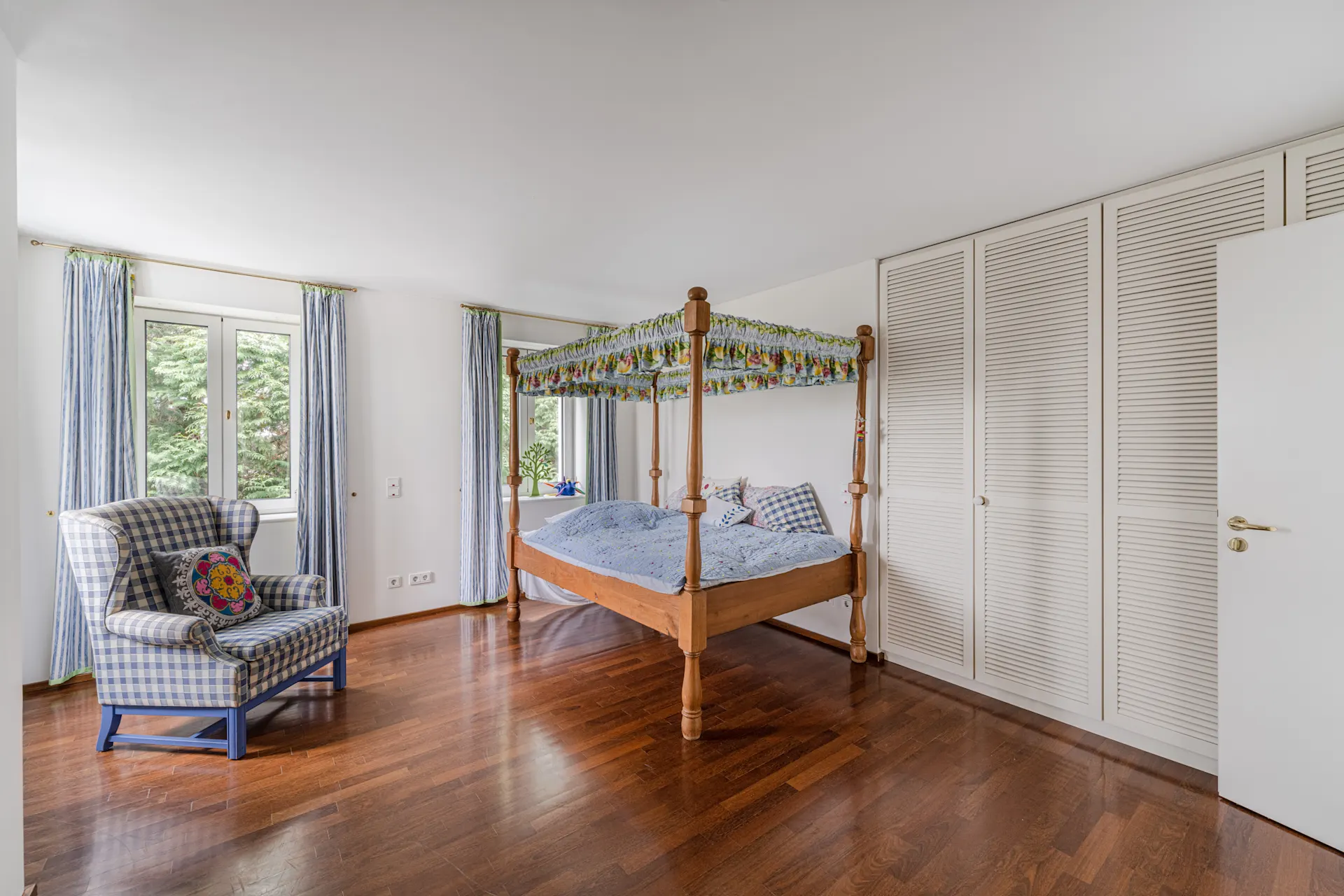 Helles Schlafzimmer mit Himmelbett, Sessel, Holzfußboden und Fenstern mit blauen Vorhängen.
