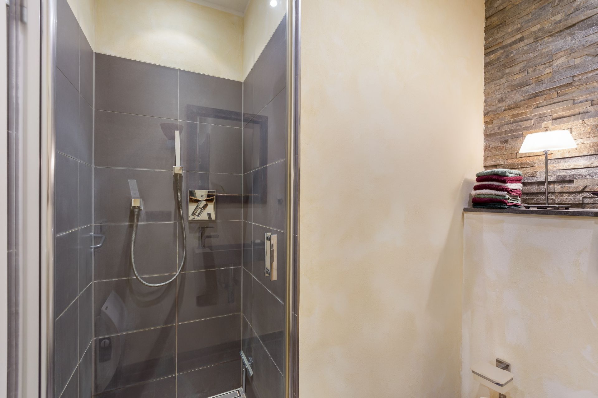 Moderne Dusche mit grauen Fliesen, daneben Ablage mit Handtüchern und Lampe.
