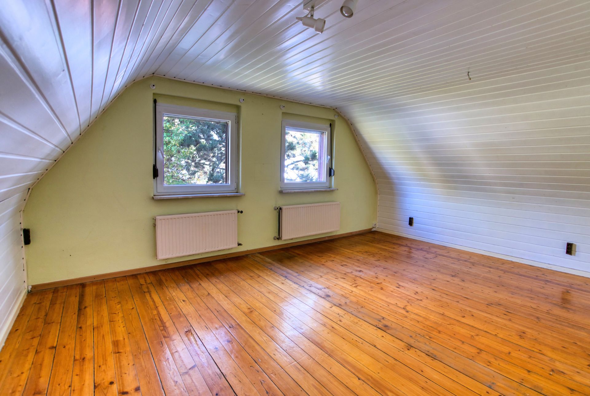 Dachzimmer mit schrägen Wänden, Holzfußboden und zwei Fenstern mit Heizkörpern.