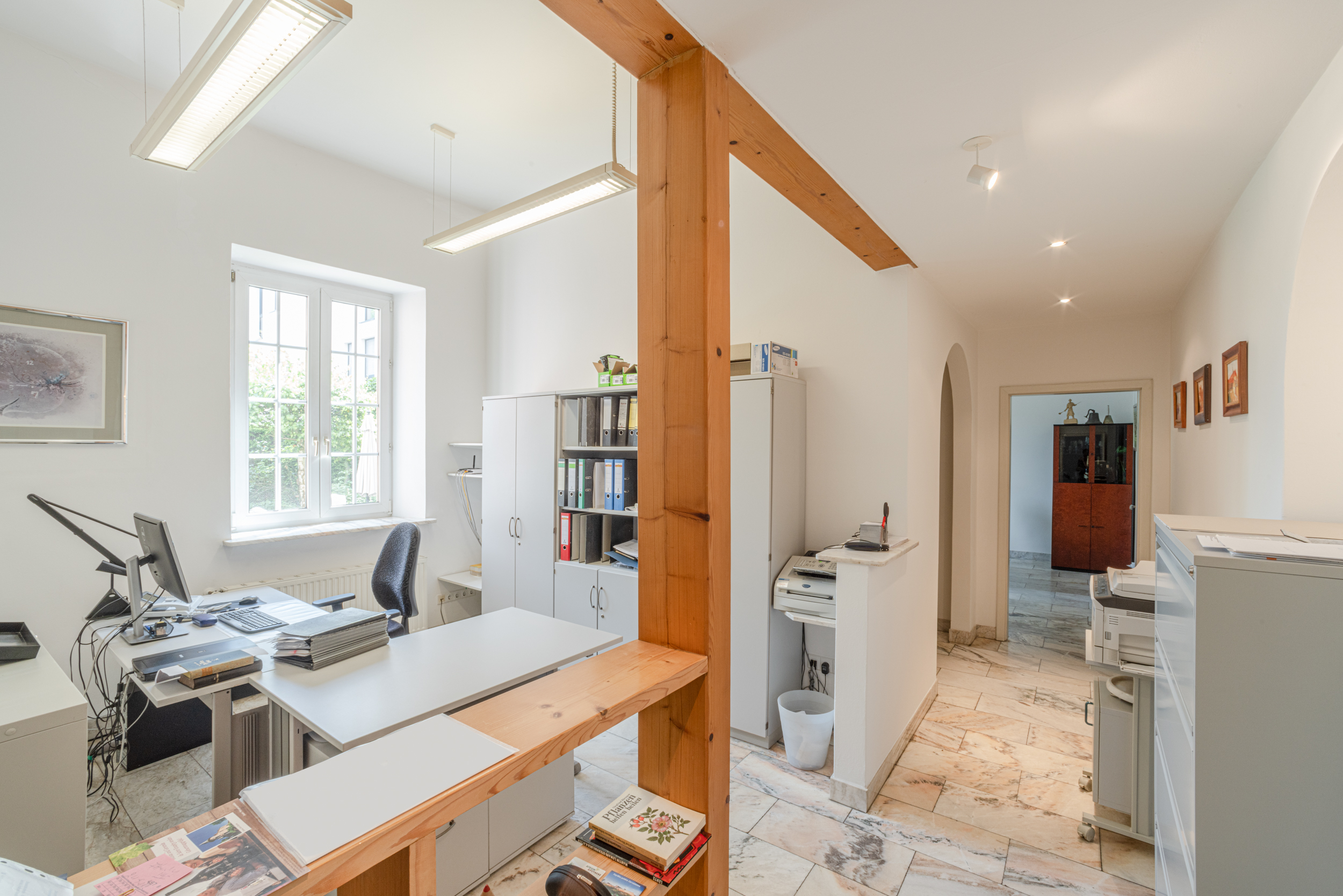 Helles Büro mit Schreibtisch, Aktenschränken, Drucker und einem Fenster, das Tageslicht hereinlässt.