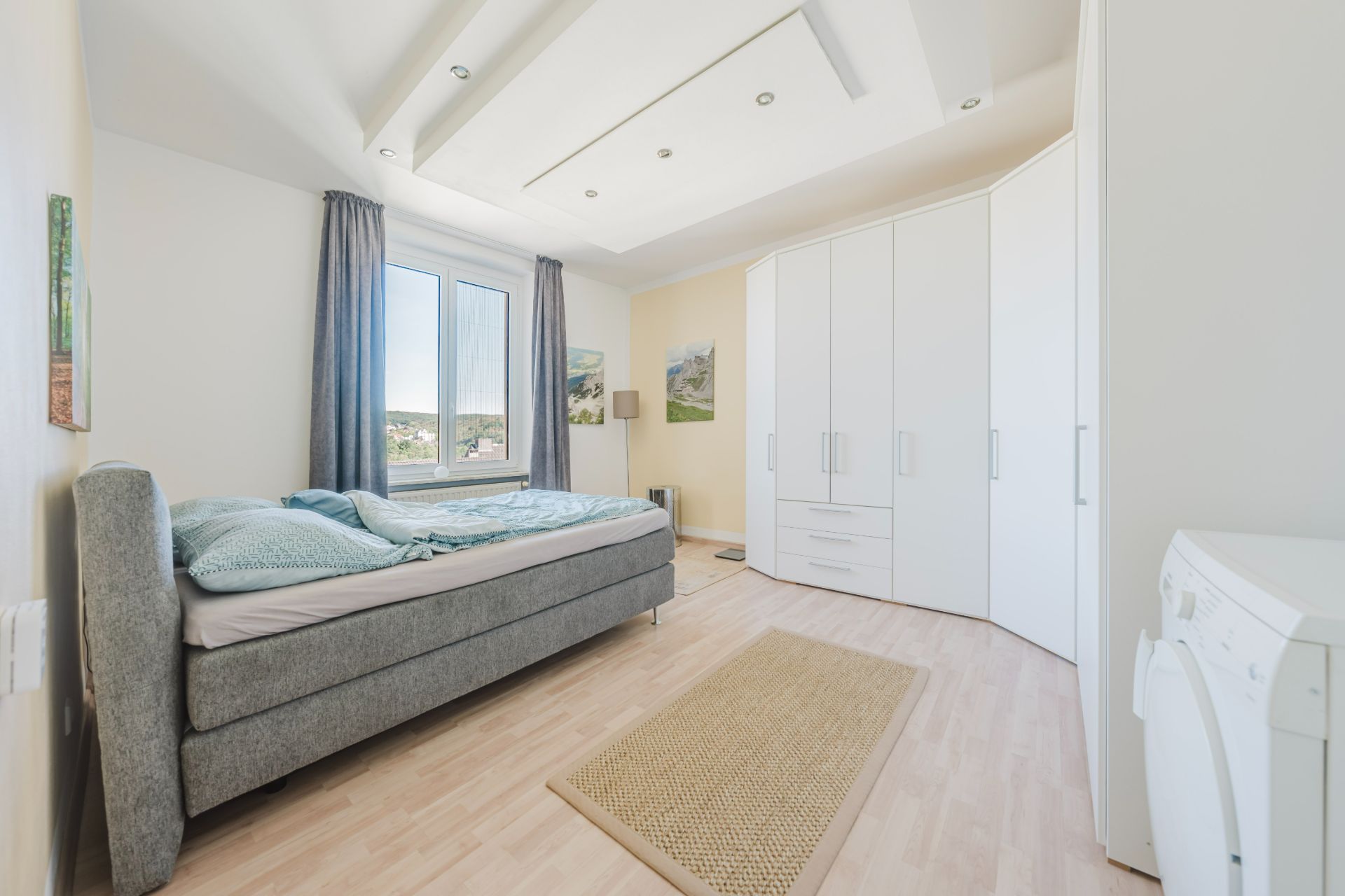 Helles Schlafzimmer mit grauem Bett, großem weißen Kleiderschrank und Fenster mit Vorhängen.