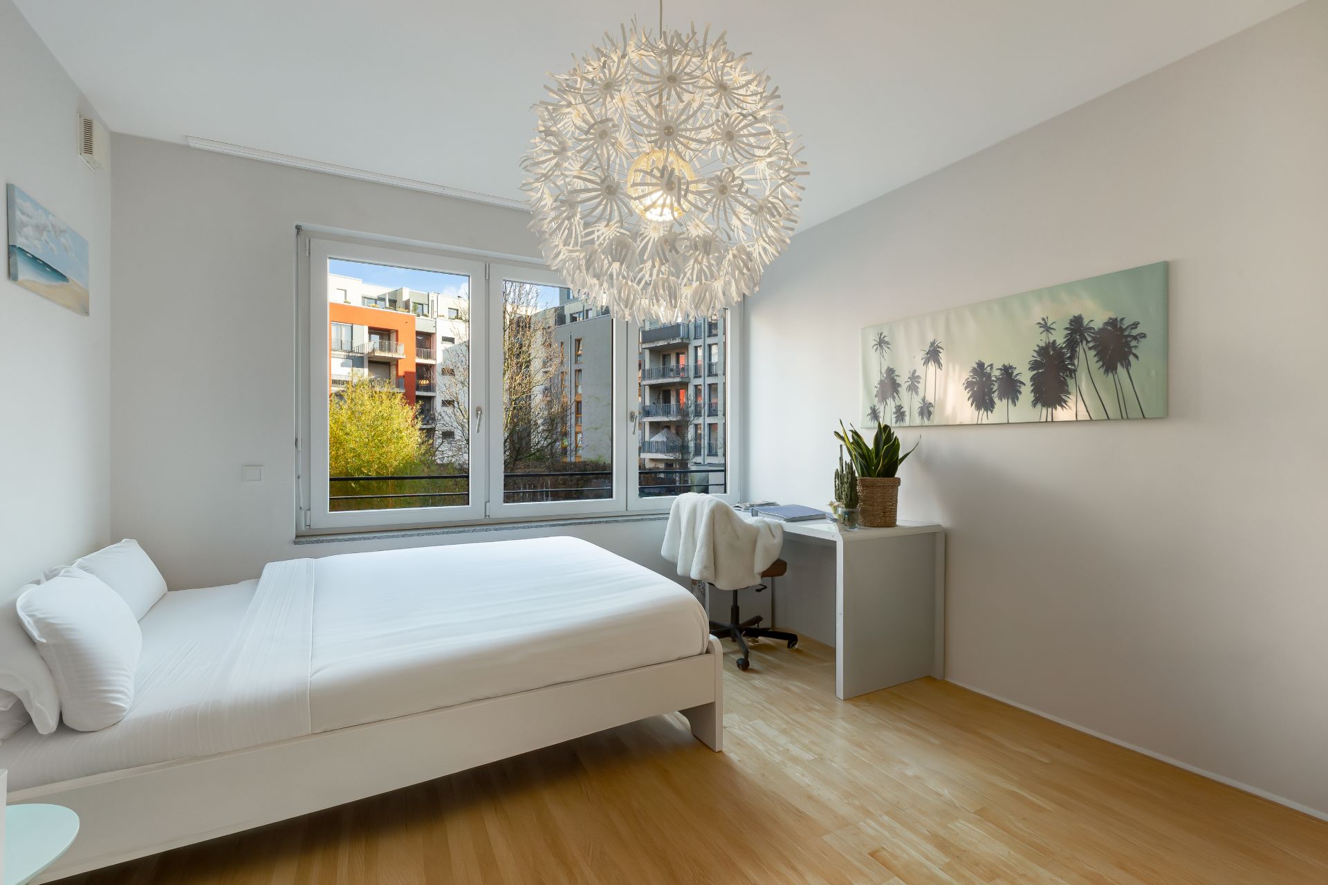 Helles Schlafzimmer mit großem Bett, Schreibtisch, Stuhl und modernem Kronleuchter vor zwei Fenstern.
