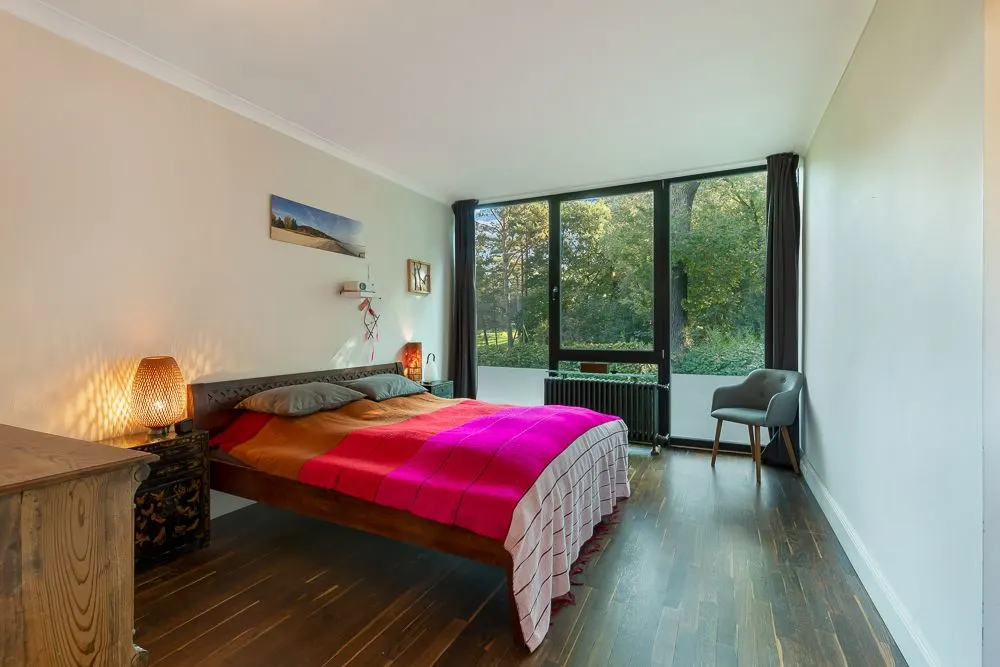 Schlafzimmer mit großem Fenster, Doppelbett und Stuhl, Holzboden und Blick ins Grüne.