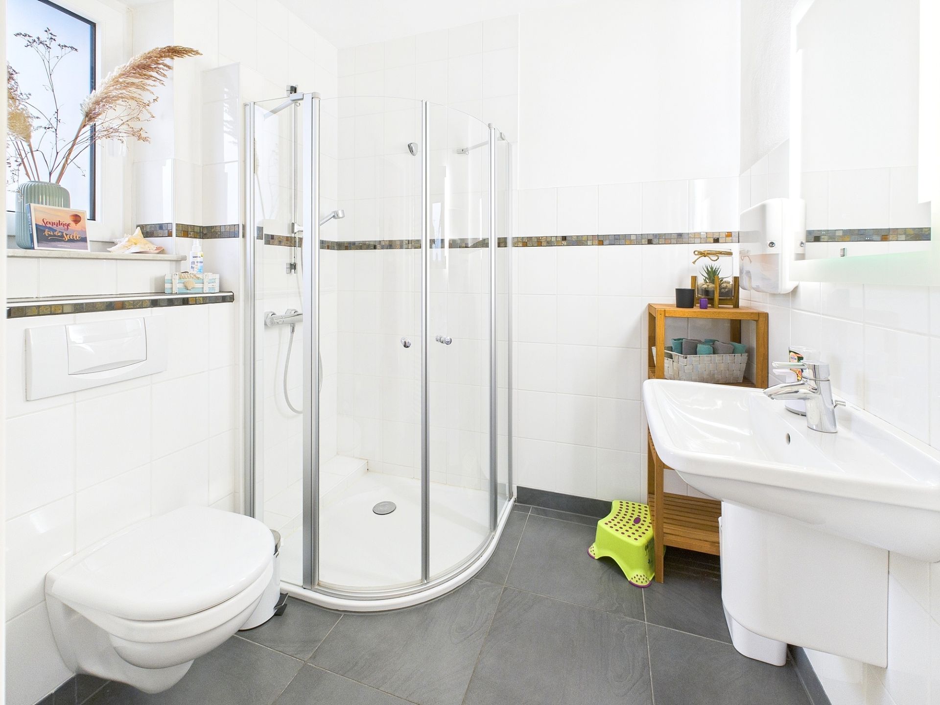 Modernes Badezimmer mit Dusche, WC, Waschbecken, Regal und Hocker auf grauen Fliesen.