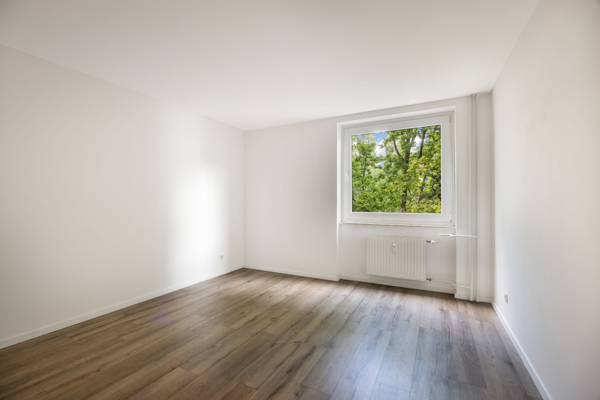 Leerer Raum mit hellem Holzboden, weißer Wand und Fenster mit Blick ins Grüne.