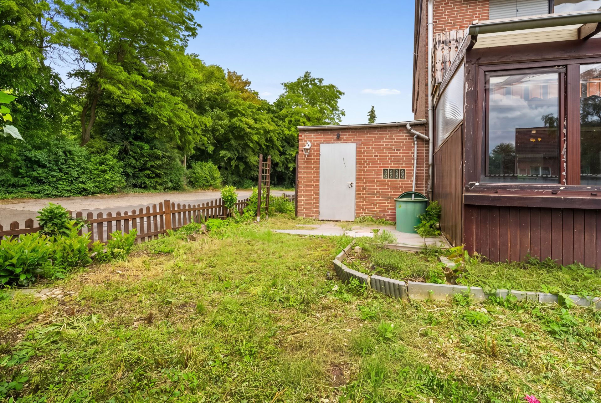 Kleiner Garten mit Rasen, Zaun, Schuppen und einem Wintergarten an einem Wohnhaus.