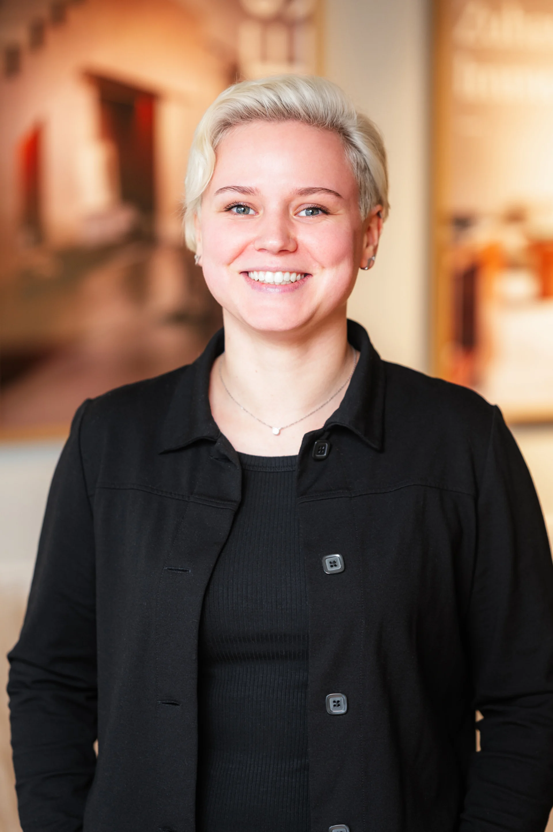 Junge Person mit kurzem, blondem Haar und blauem Oberteil steht in einem modernen Innenraum.