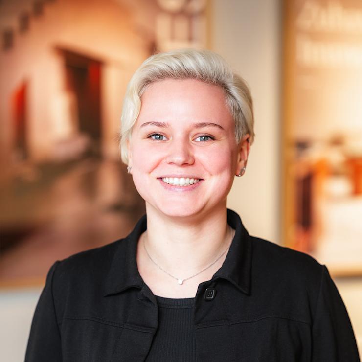 Junge Person mit kurzem, blondem Haar und blauem Oberteil steht in einem modernen Innenraum.