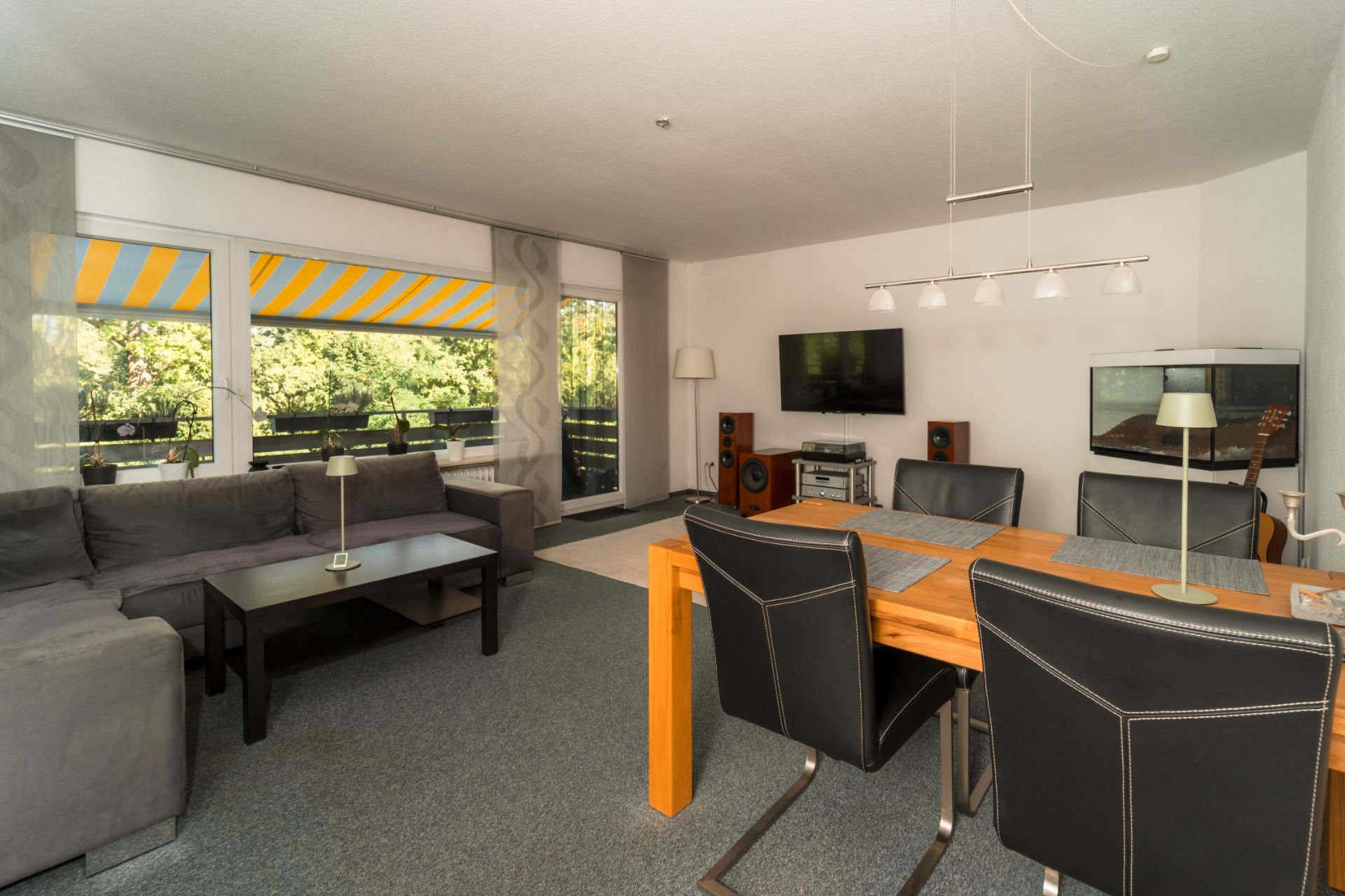 Wohnzimmer mit grauem Sofa, Esstisch, TV, Aquarium und großen Fenstern mit gelber Markise.