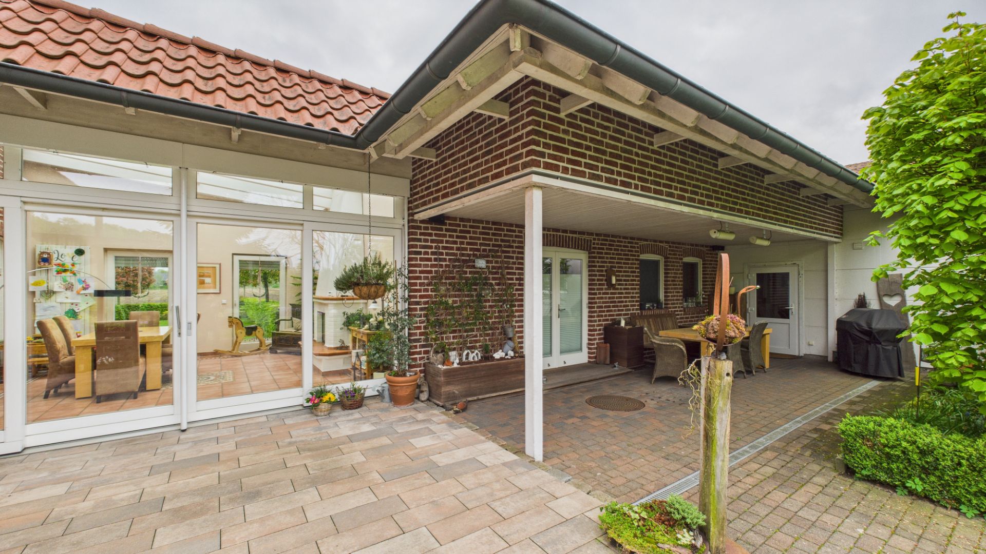Gepflasterte Terrasse mit Überdachung, Gartenmöbeln und viel Glasfassade an einem Wohnhaus.